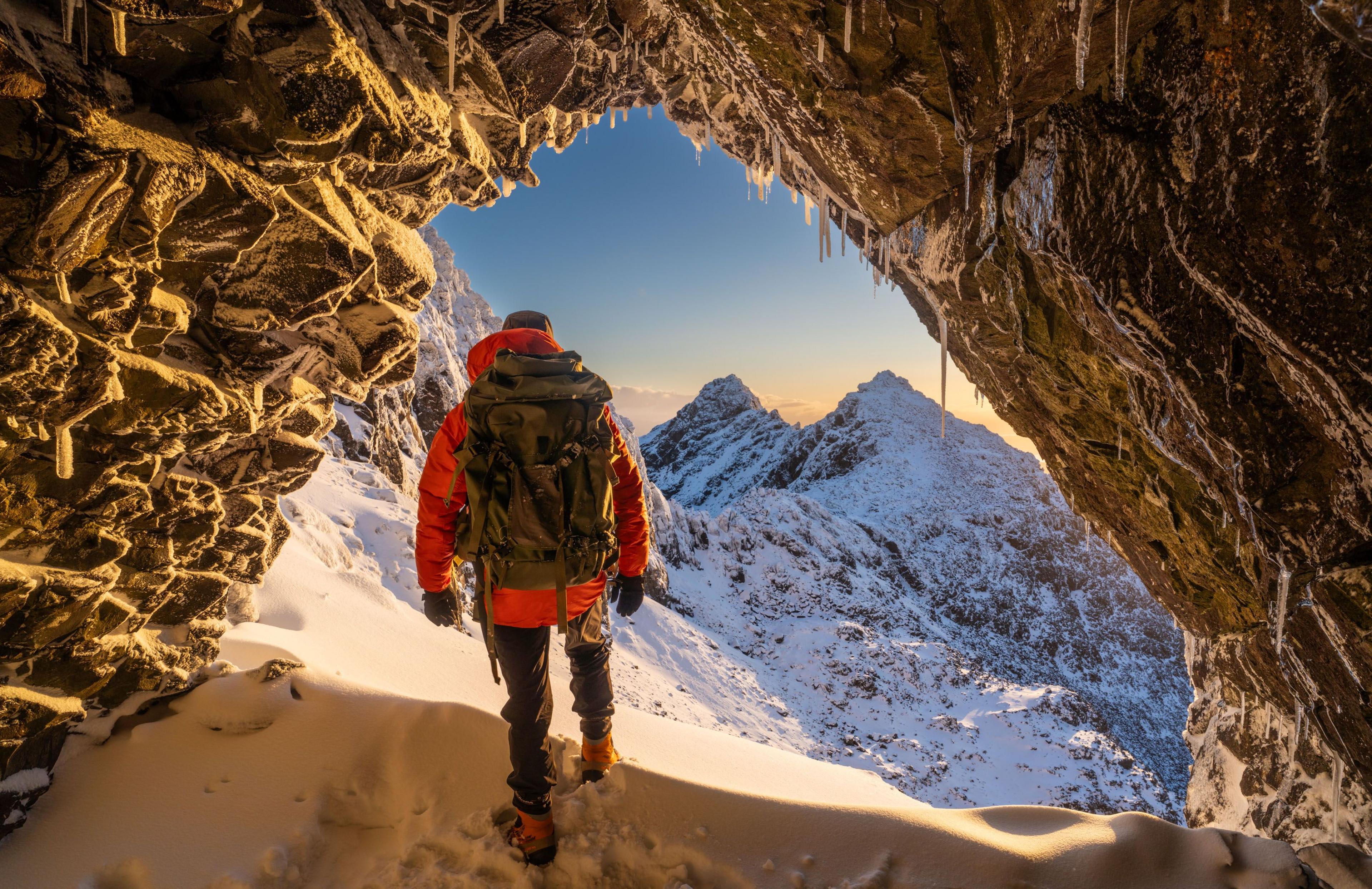 A man in mountain climbing gear with a large backpack walks out of a cave into a snow-capped set of peaks