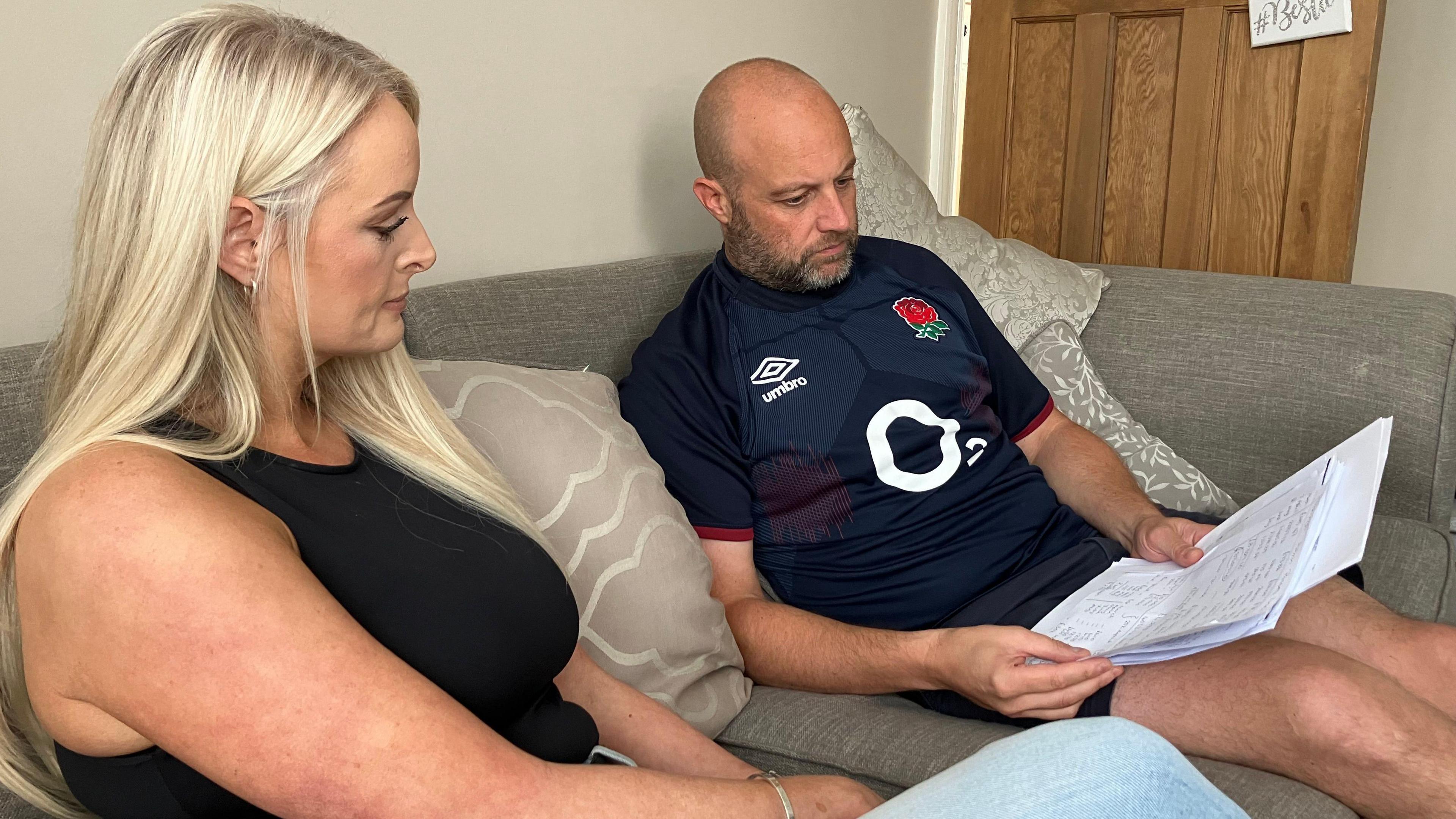 Emma and James Richardson sit on a grey sofa in their home. James is holding a handful of documents, which they are both looking at.