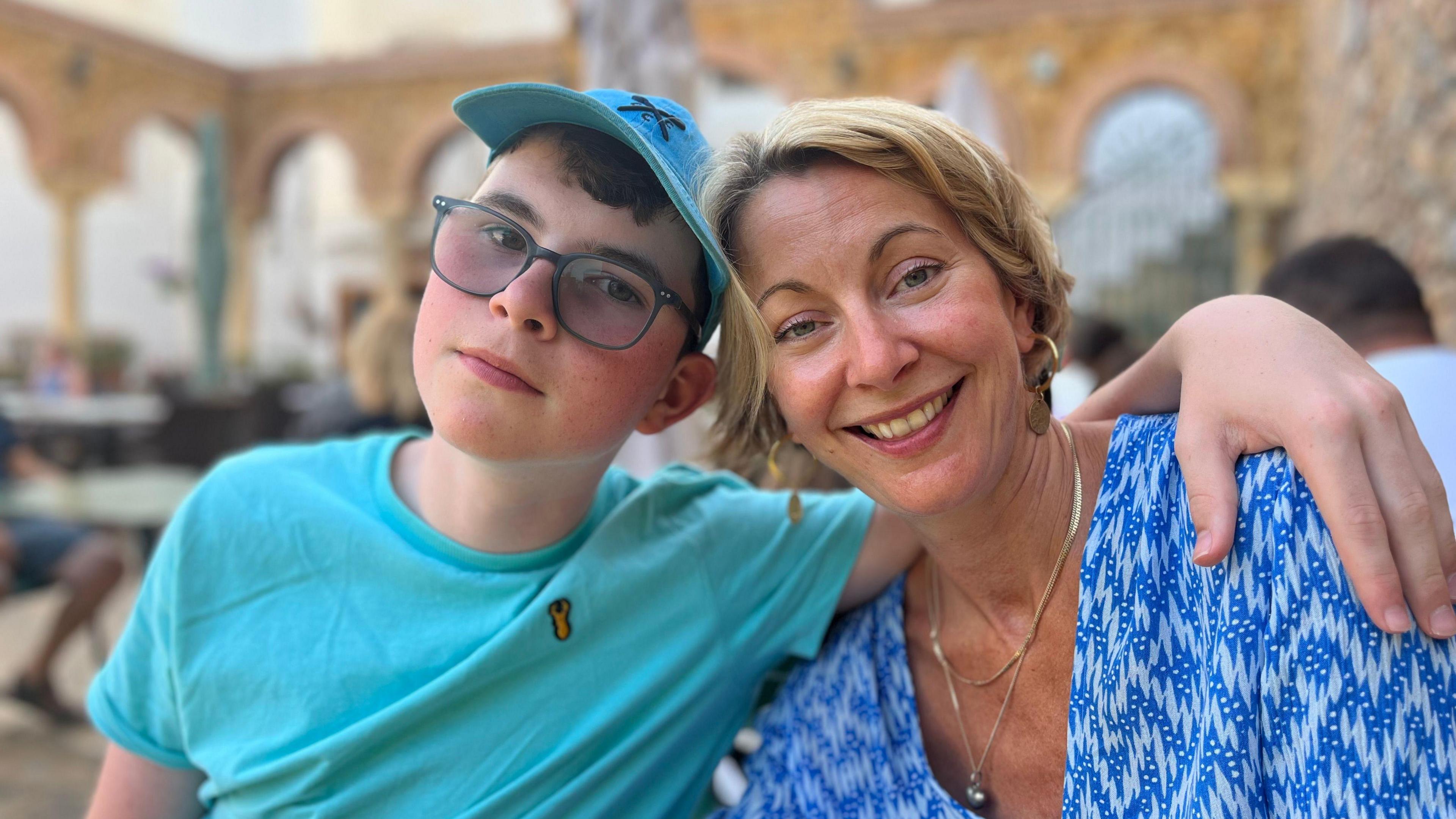 Teenage boy, Lucas, wearing a blue t-shirt, blue hat and black glasses with his arm around his mum, Louise wearing a blue and white blouse.