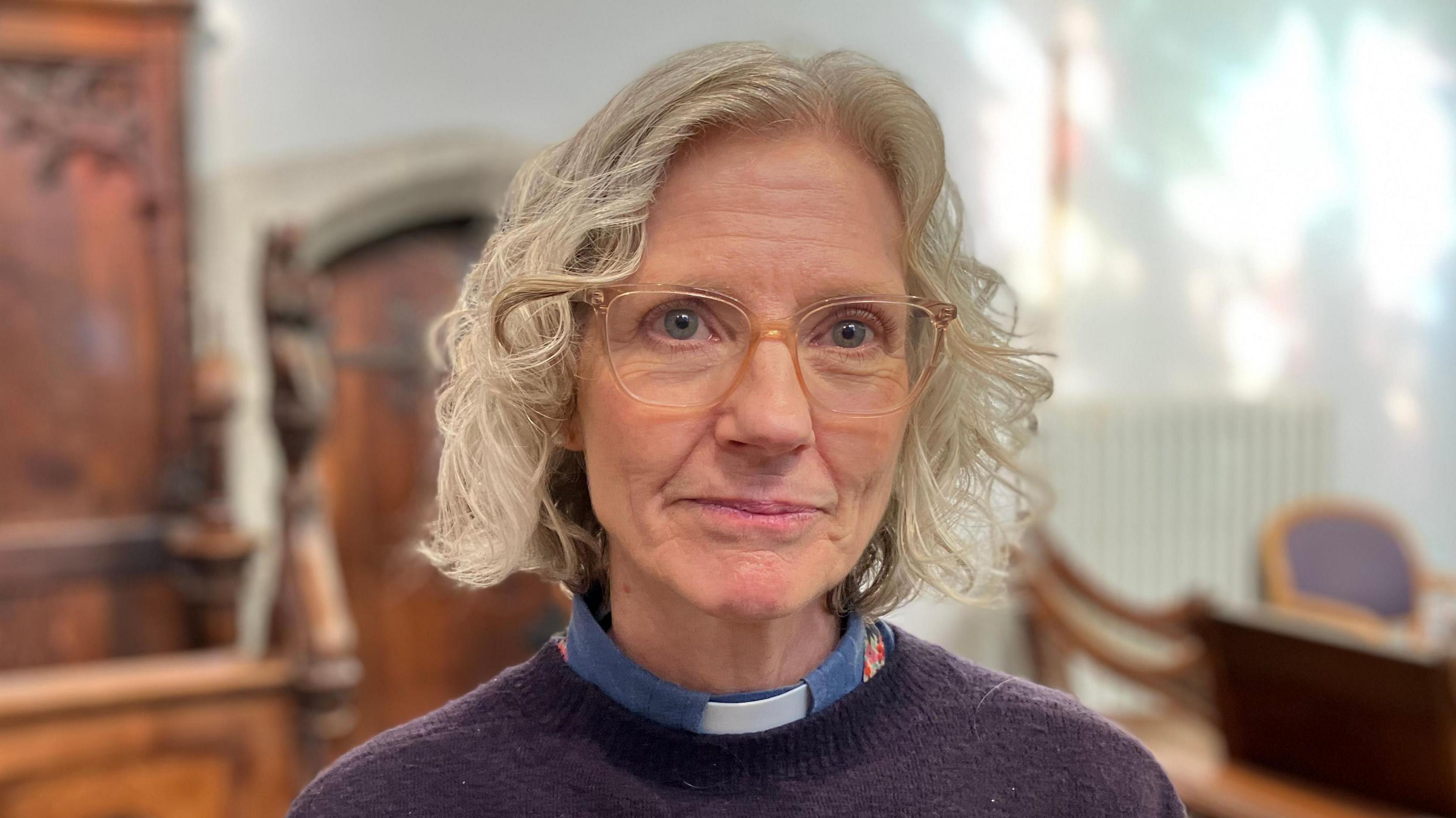 Reverend Jane Smith is in the middle of the image. She has short blonde curly hair and is wearing clear framed glasses, a purple jumper and blue dog collar. 