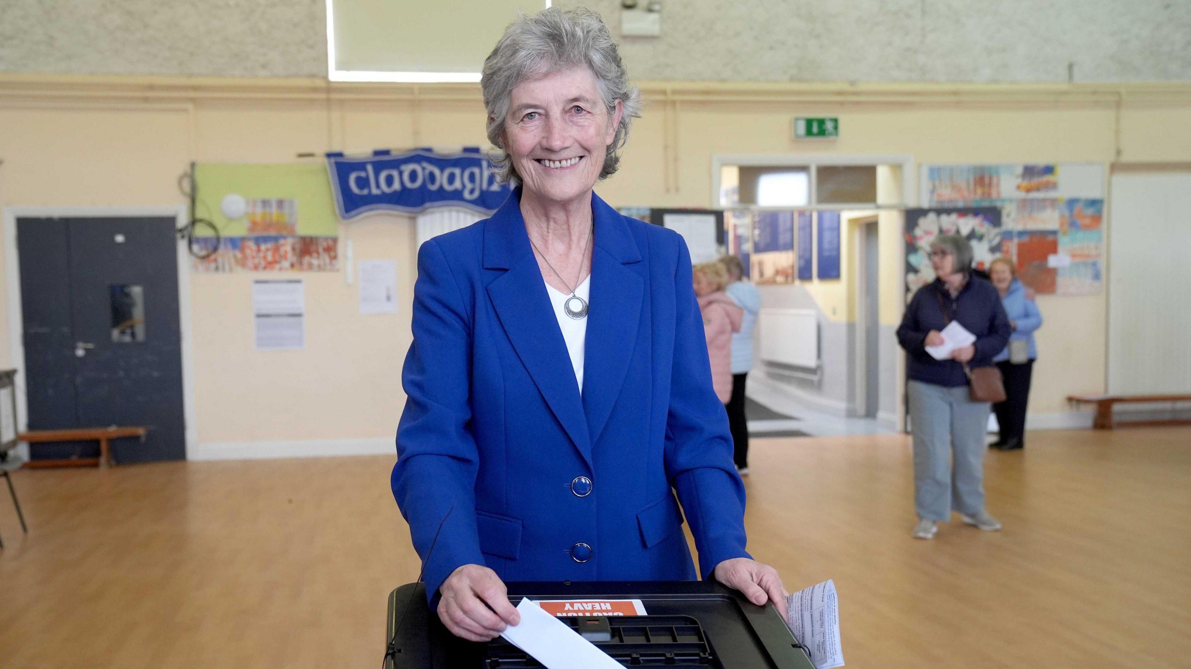 A woman with short grey hair stands at a black box, smiling at the camera. She is holding a piece of paper, which she is putting into the box. She is wearing a blue blazer and white shirt.