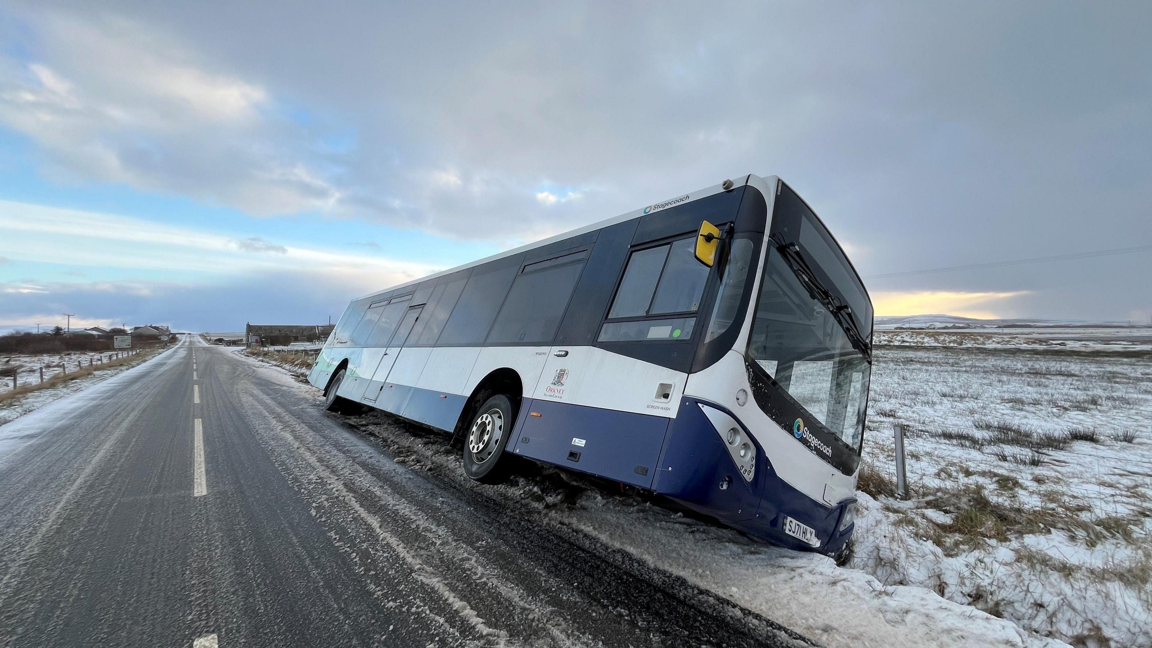 A bus leans in a roadside ditch, surrounded by snow and ice 