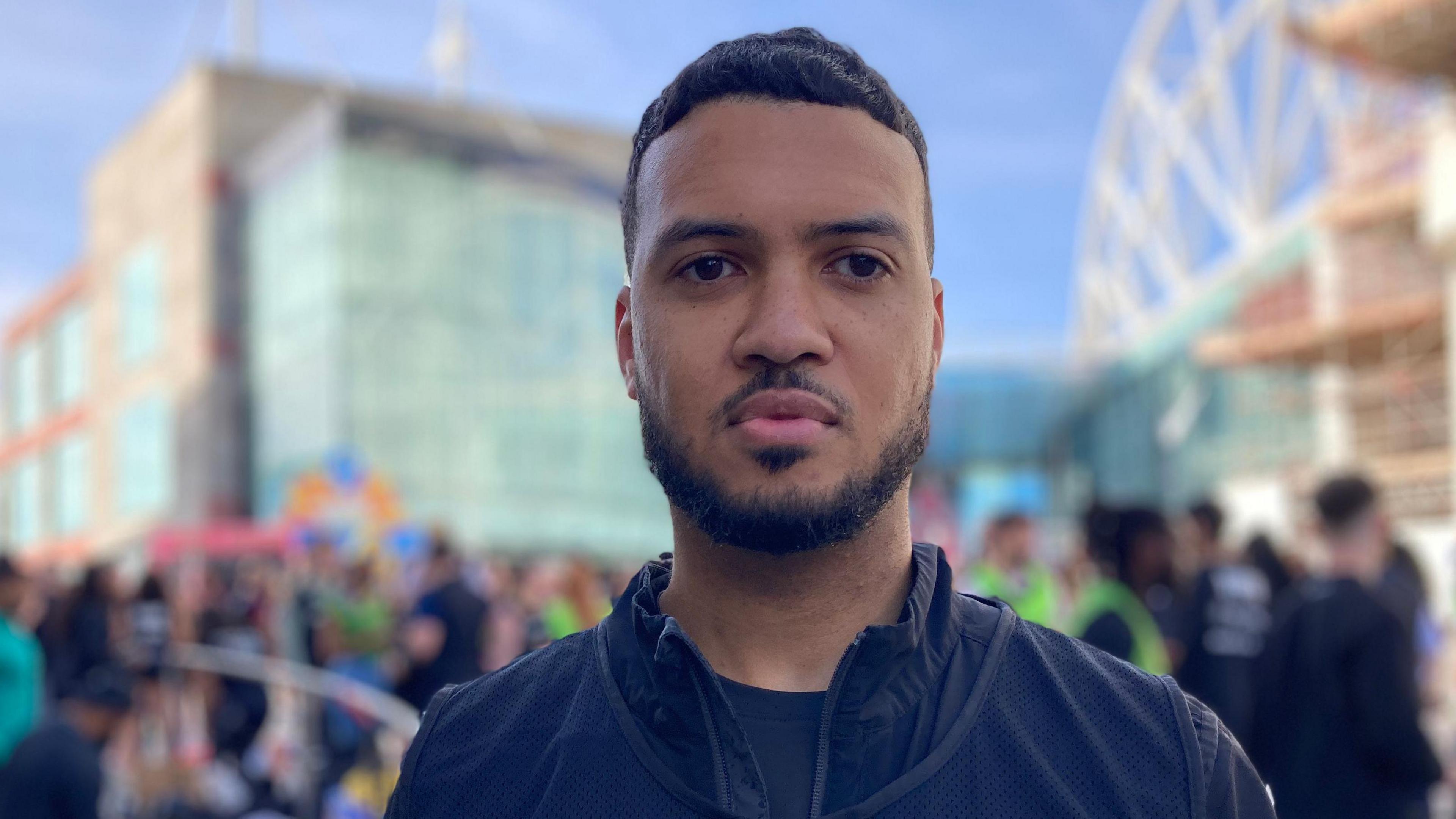 Jonathan Sillwood is looking out of shot while standing in Birmingham city centre. He has short dark hair and similar coloured beard. He is wearing a dark blue gym top