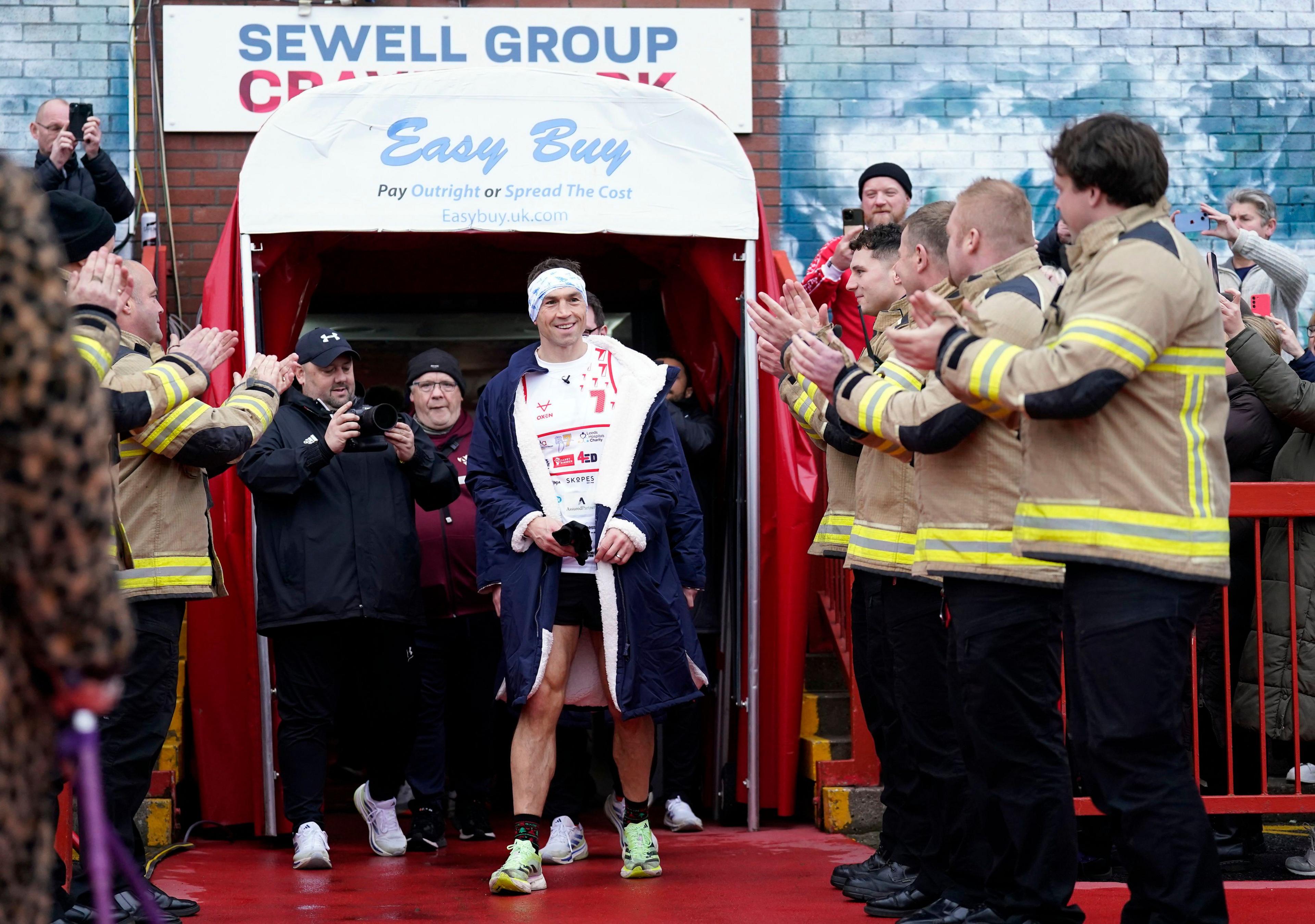 Two lines of firemen clap and cheer for Kevin Sinfield as he arrives at Craven Park through the player's entrance. Crowds of people either side cheer and take photos. Mr Sinfield is smiling.