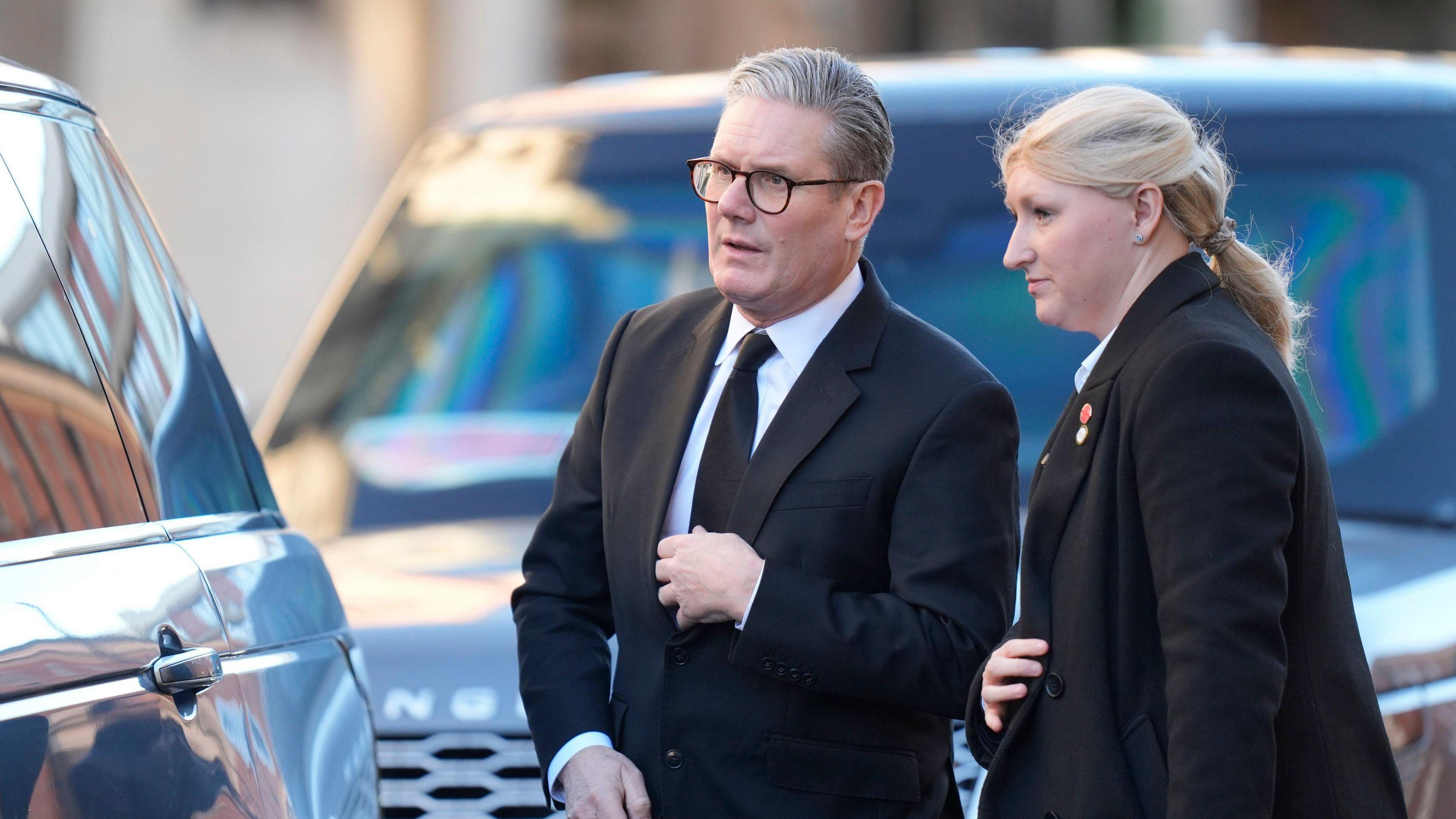 Prime Minister Sir Keir Starmer in a black suit and tie. He is wearing glasses and has grey hair and is standing next to a blonde woman wearing a black blazer. They are next to two Range Rovers