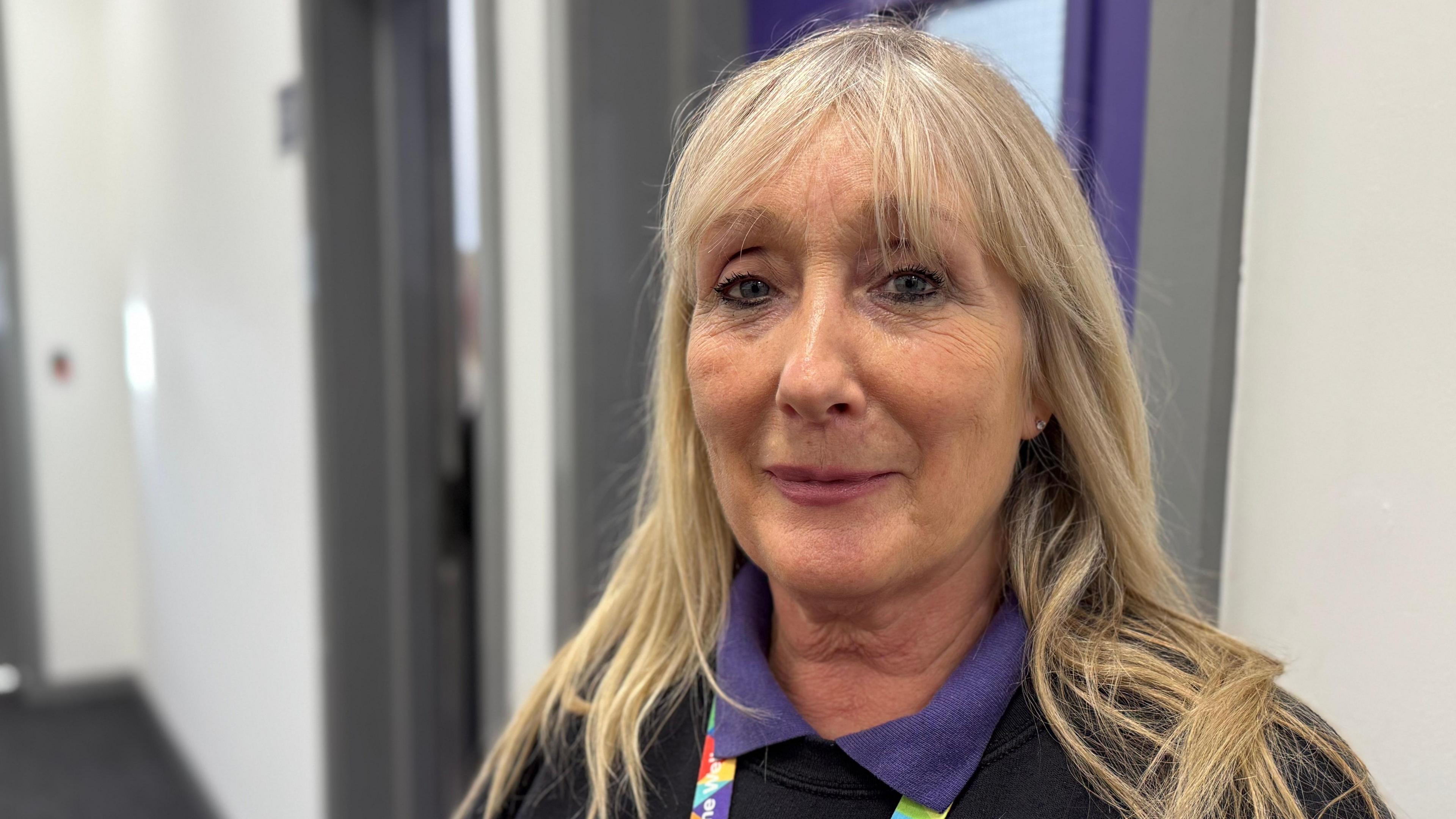 A woman with blonde hair just beyond the shoulders in length wearing a black jumper over a purple polo t-shirt. She has a lanyard around her neck and is standing inside a school classroom.