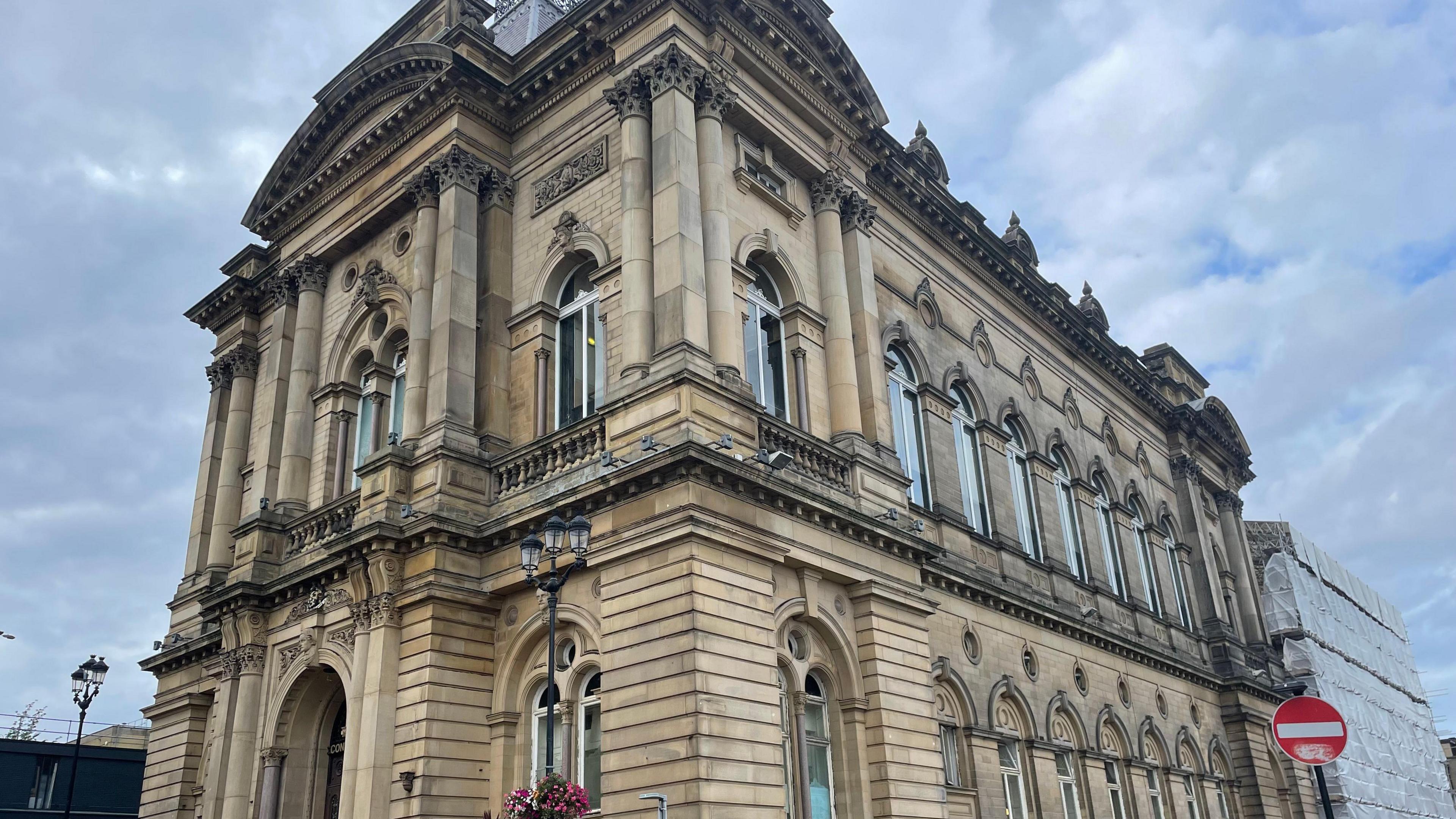 Huddersfield Town Hall, which is a Grade II-listed building, with clouds gathering overhead. The back right hand side of the building has building work taking place, with scaffolding in place.