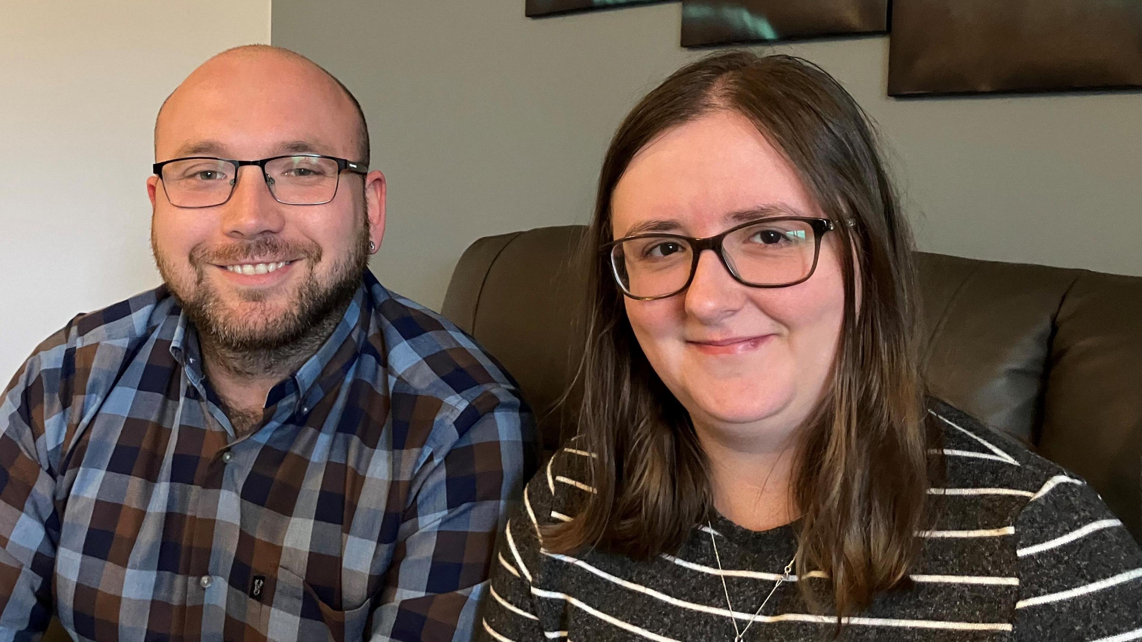 A balding man with a beard and glasses sitting on a sofa at home next to a woman with long brown hair and glasses