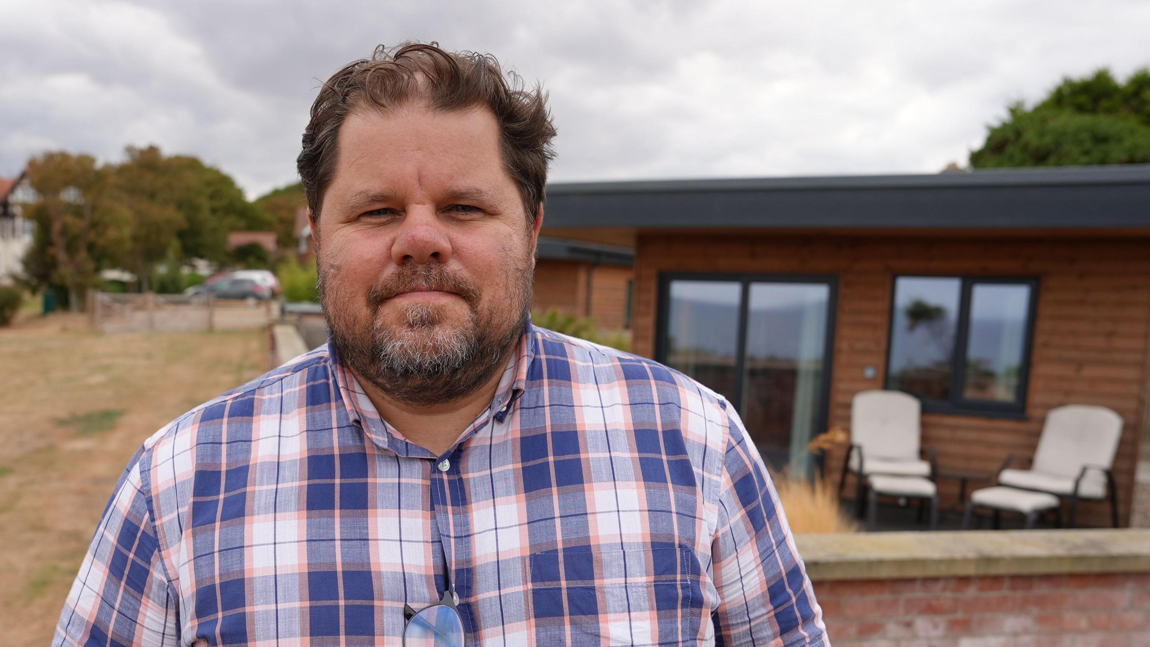 Alex Ponting, a man standing outside the Sea Marge Hotel in Overstrand near Cromer. Behind him is a wooden mobile lodge. He is looking directly at the camera and is wearing a blue, white and orange checked shirt.