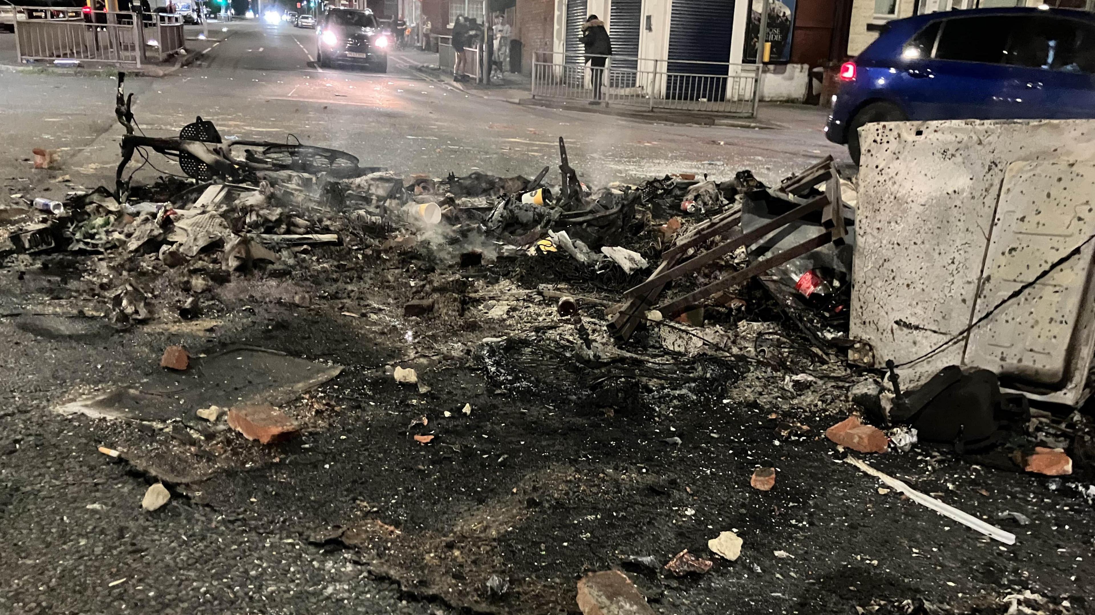 The embers of some heavily burnt property on a public street. A bike can be seen abandoned at the back of the rubble and in the far background is a car with its headlights on.