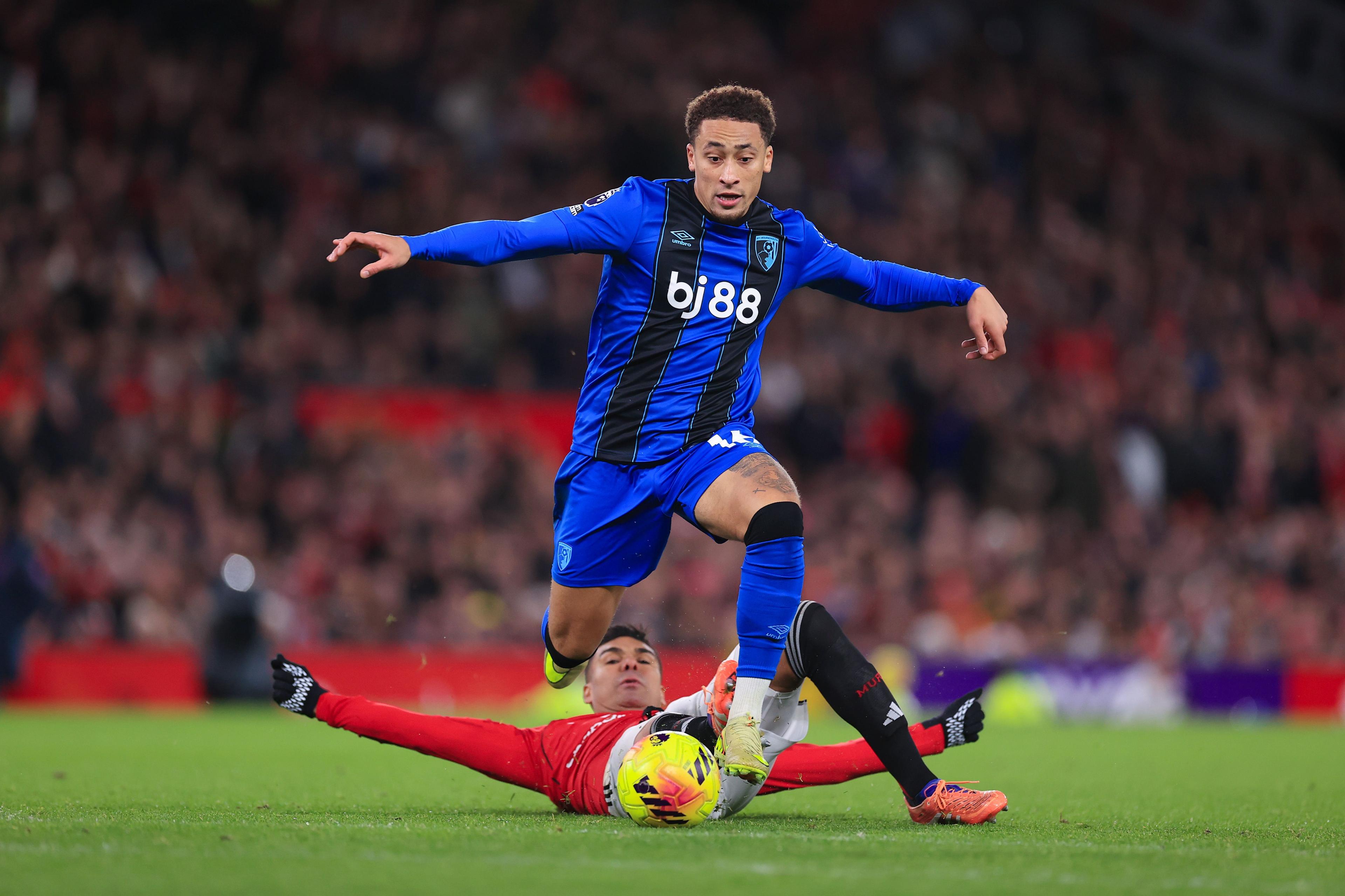 A football player in a blue and black kit sprints forward while an opponent in red slides in from the ground, attempting to tackle the ball. The action takes place on a brightly lit pitch with a packed stadium crowd blurred in the background.