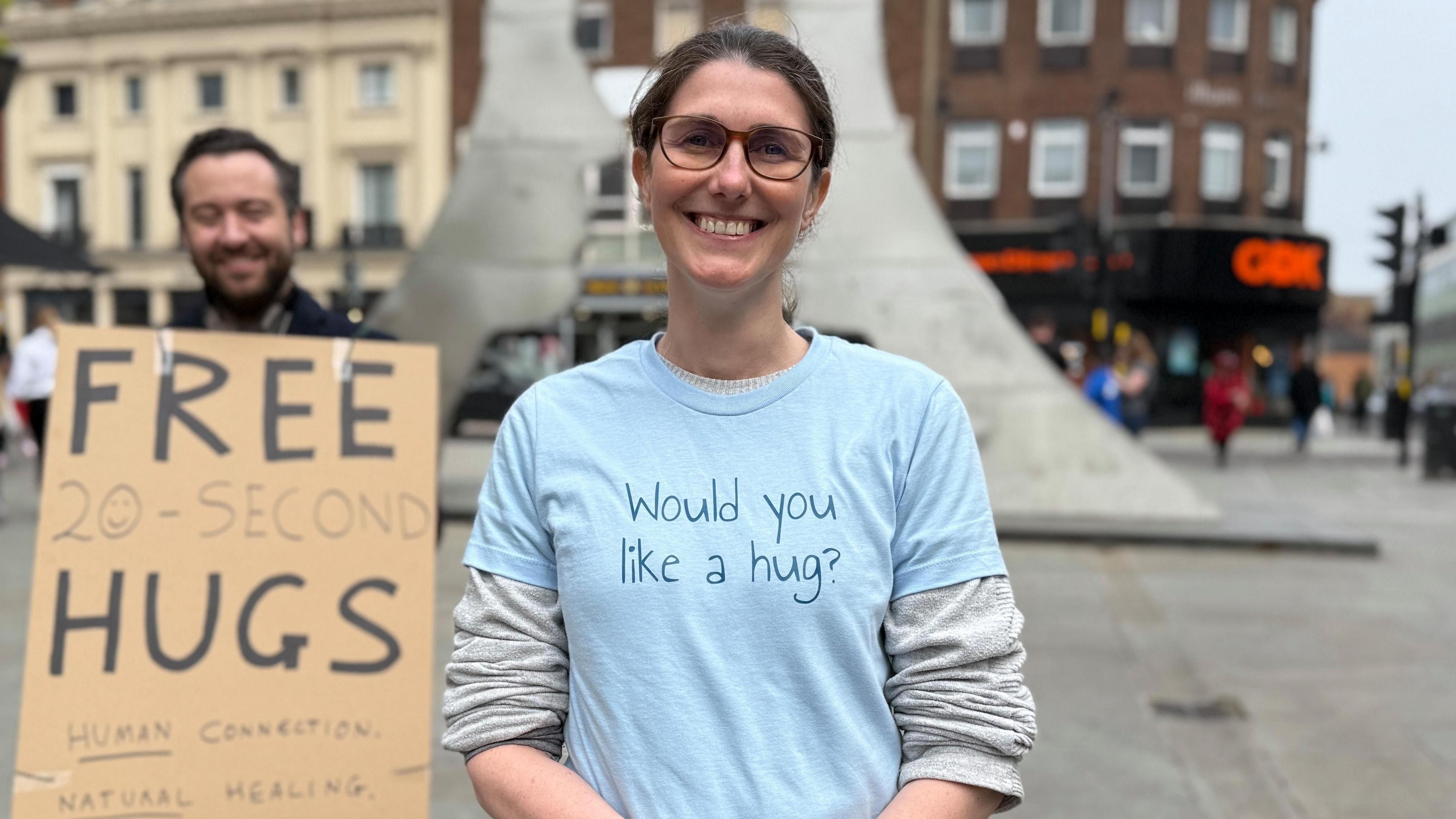 A smiling woman wearing glasses and a blue t-shirt with the words "Would you like a hug?" stands in the street in front of a man, who is also smiling, holding a sign that reads "FREE 20-SECOND HUGS".
