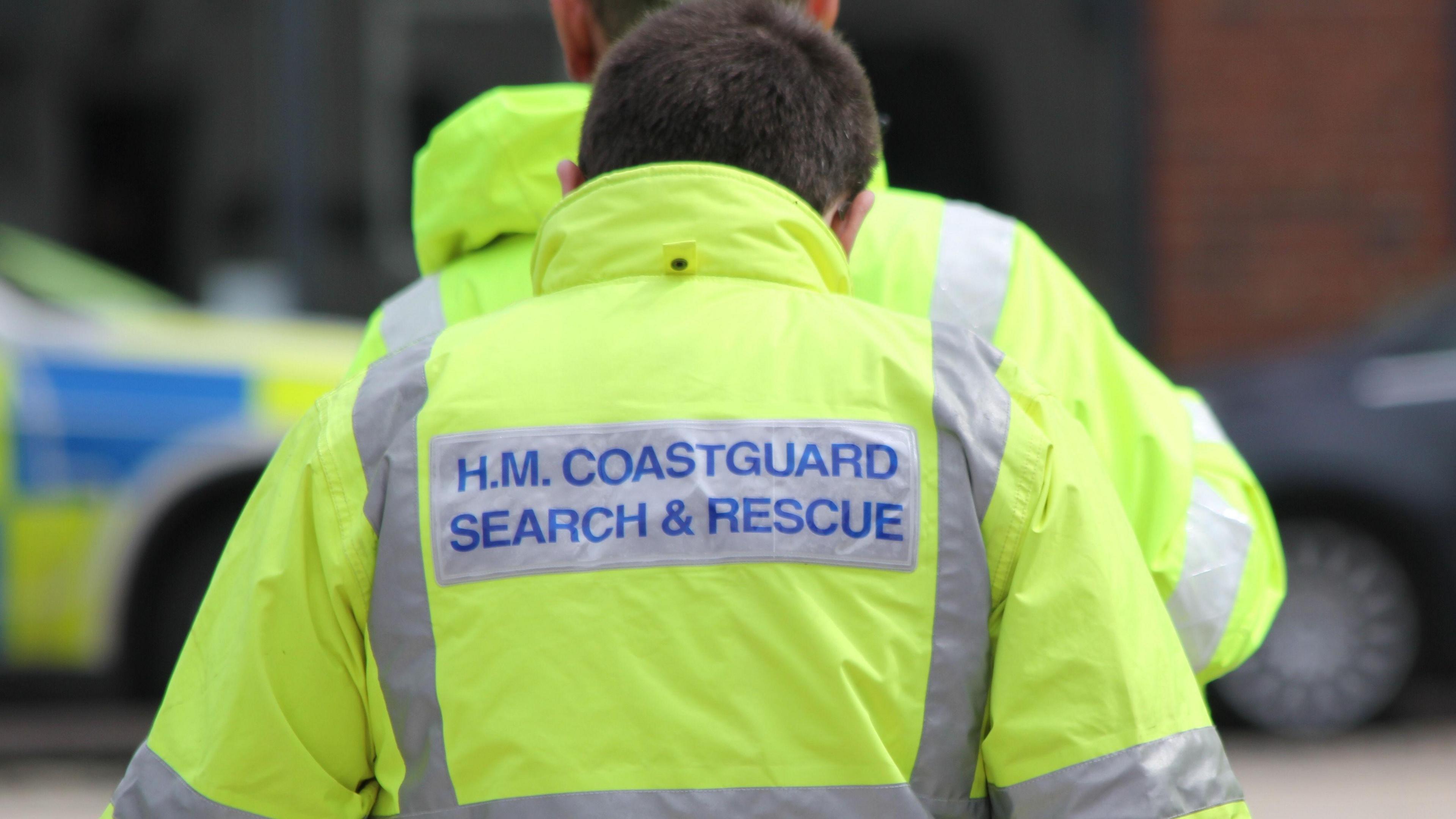 The image shows the back of a coastguard wearing a fluorescent jacket with the word HM Coastguard Search and Rescue on it.