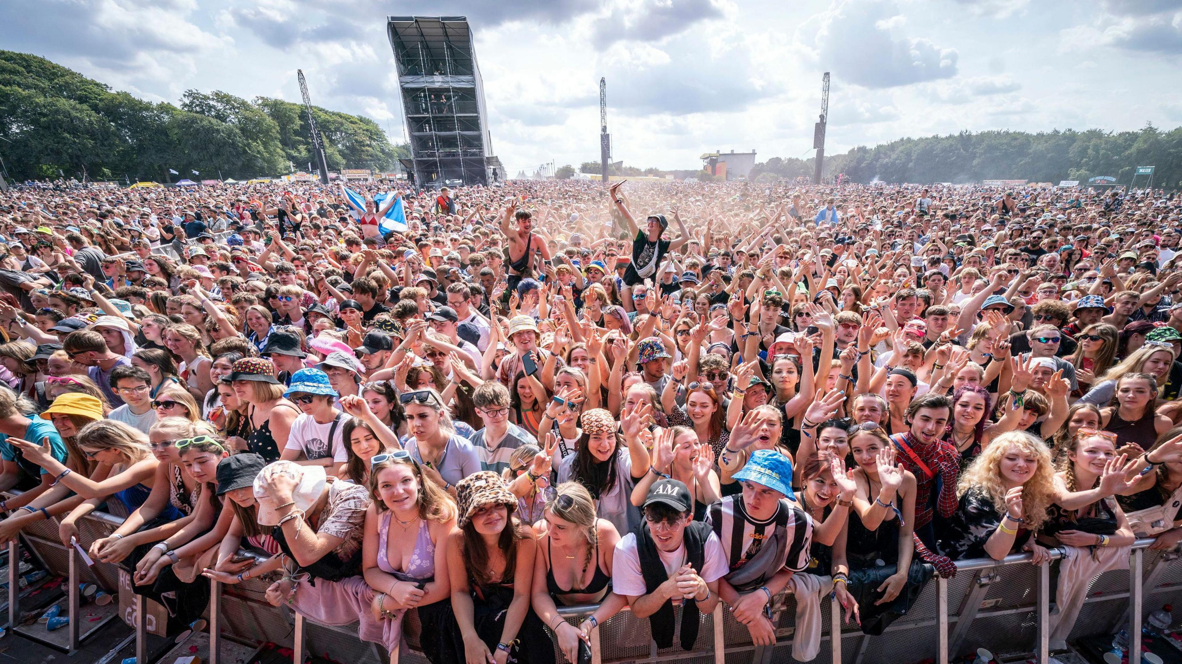 A large crowd of people in summer clothes, as seen from just in front of a stage. People at the front have their arms resting on a barrier and some are waving towards the camera. A scaffolding tower can be seen further back in the crowd and trees surround the site.