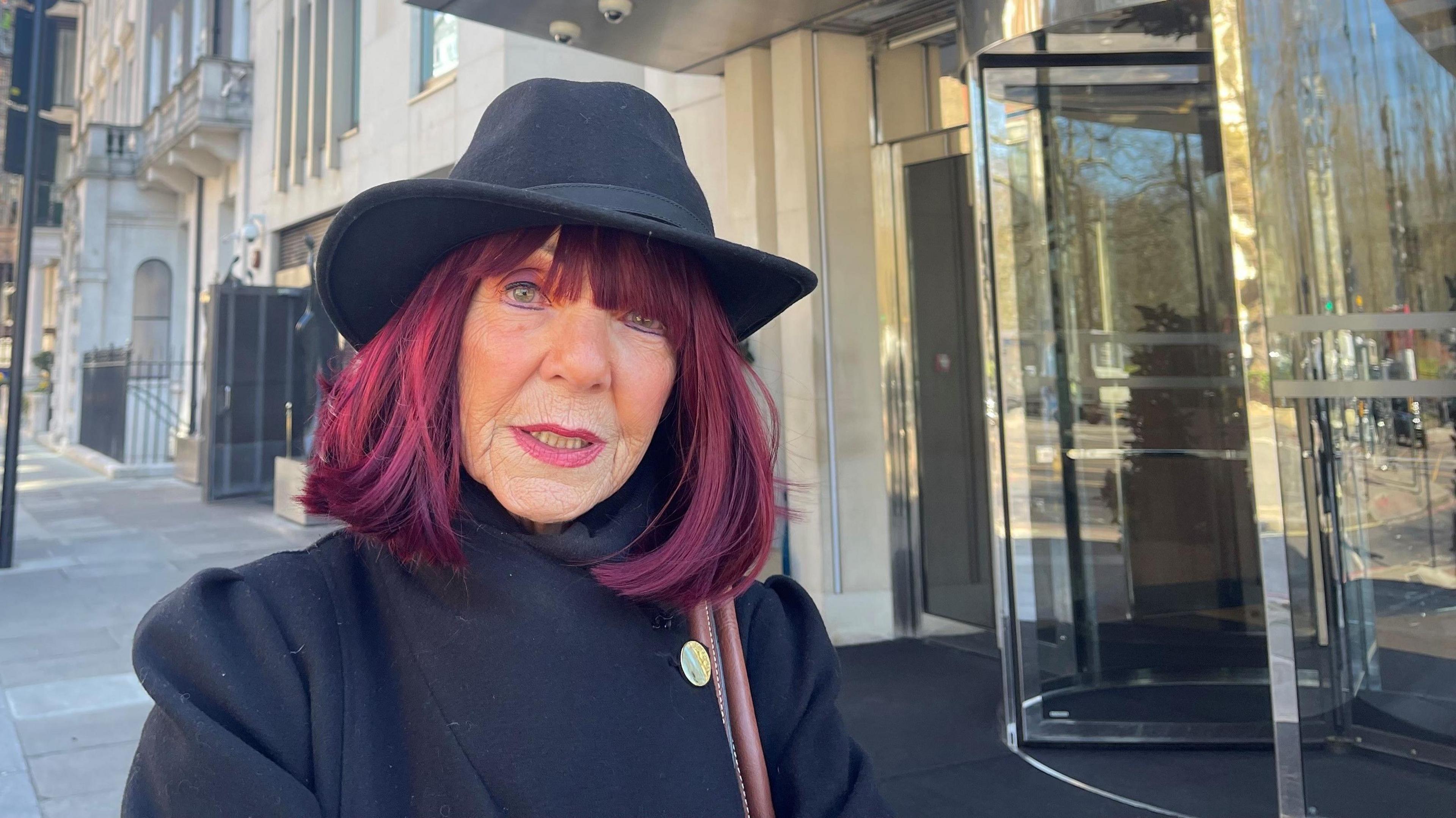 Mature woman with dyed red hair standing on a pavement next to a building. She wears a black coat and black hat and looks quizzical.