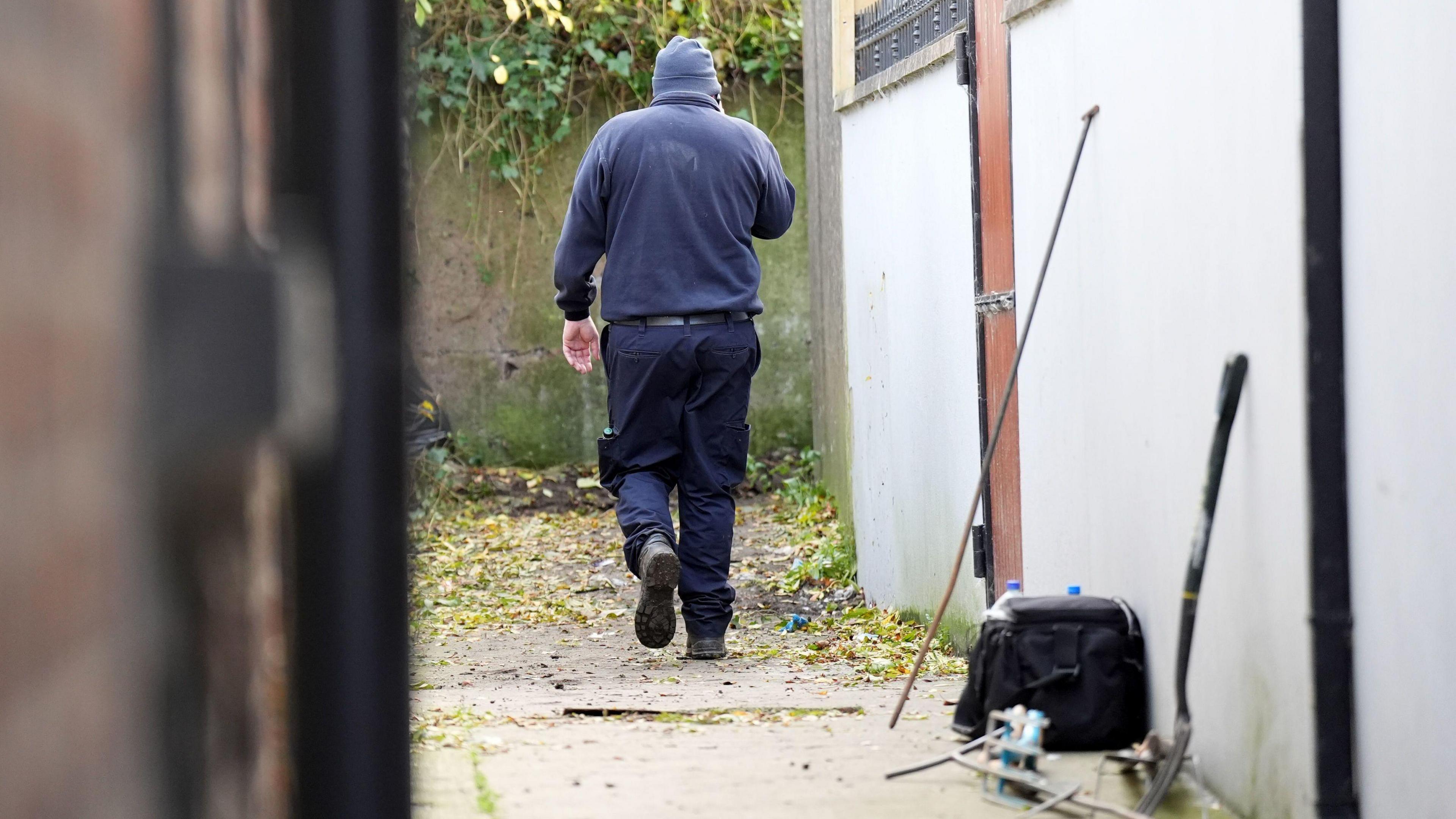 A member of the garda team involved in the search for Kyran Durnin.  The man is wearing dark blue clothing, boots and a beanie hat.   He is walking down an alleyway towards an ivy-covered wall. There is a gardening fork, a bag and bottles in the foreground.