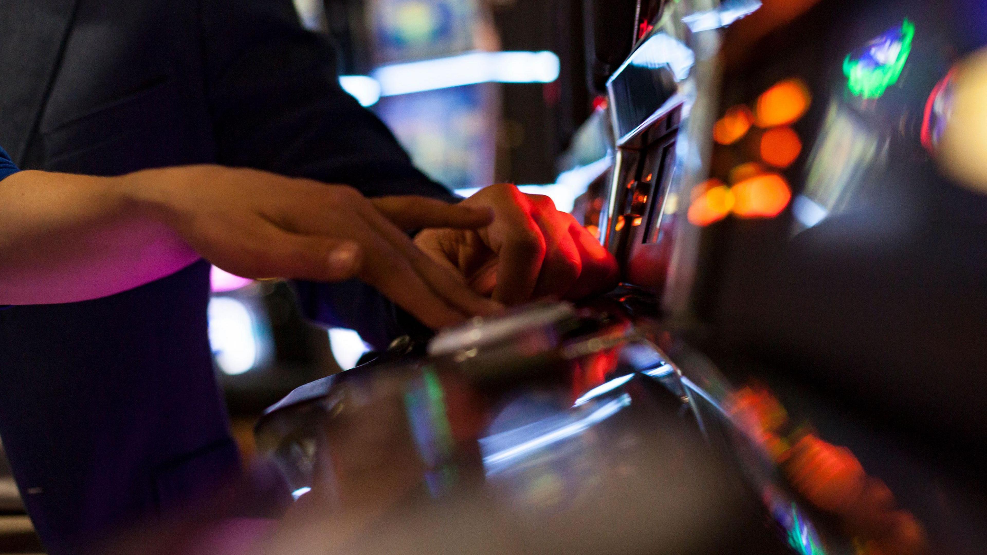 Close up shot of two hands (from two people) at a slot machine. You can see some out of focus orange spots of light and then some purple neon reflecting on the arm of the first person in the foreground.
