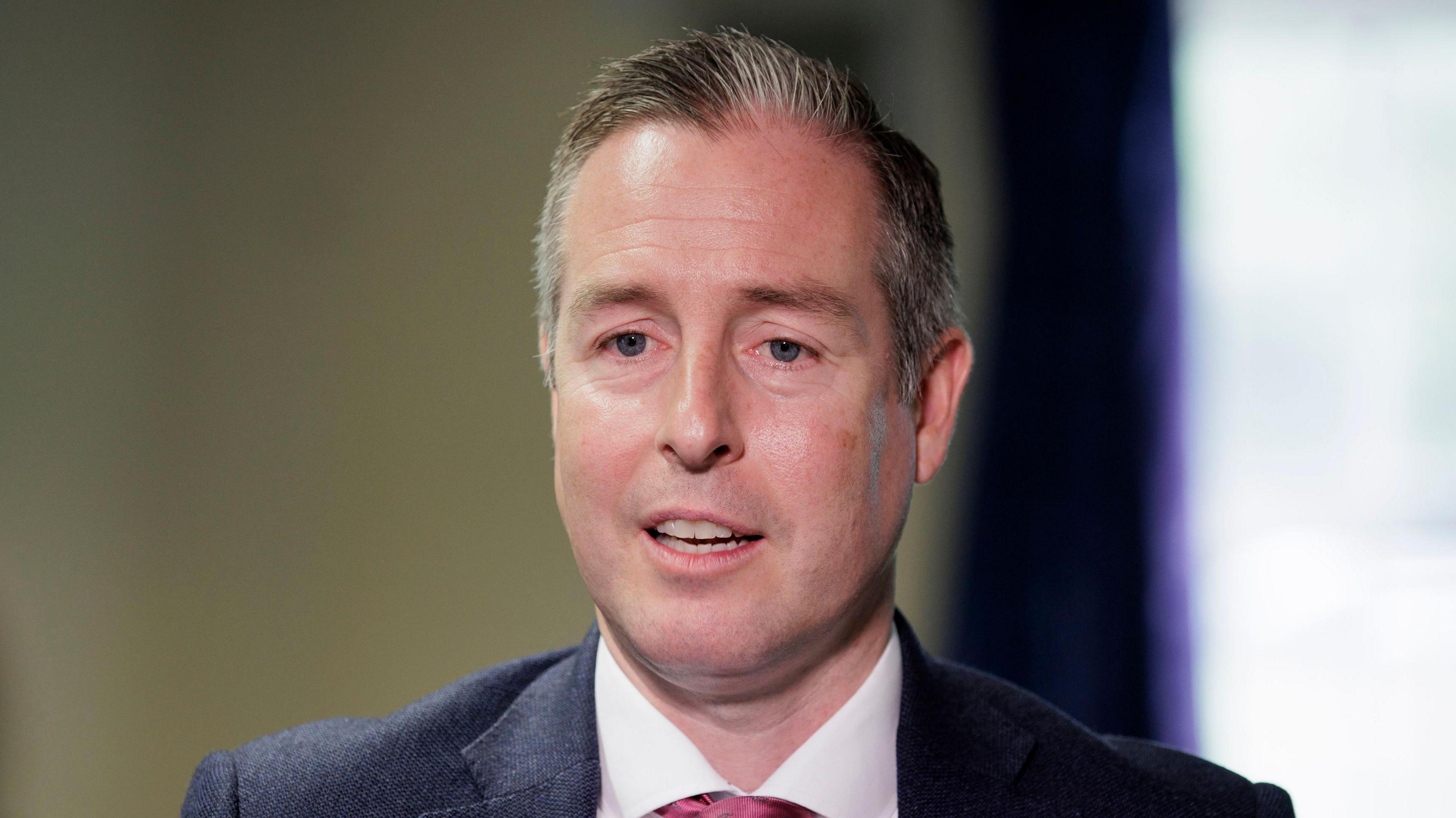 The Education Minister, Paul Givan pictured wearing a dark coloured suit and a red tie. Behind his is a magnolia coloured wall and purple curtains. 