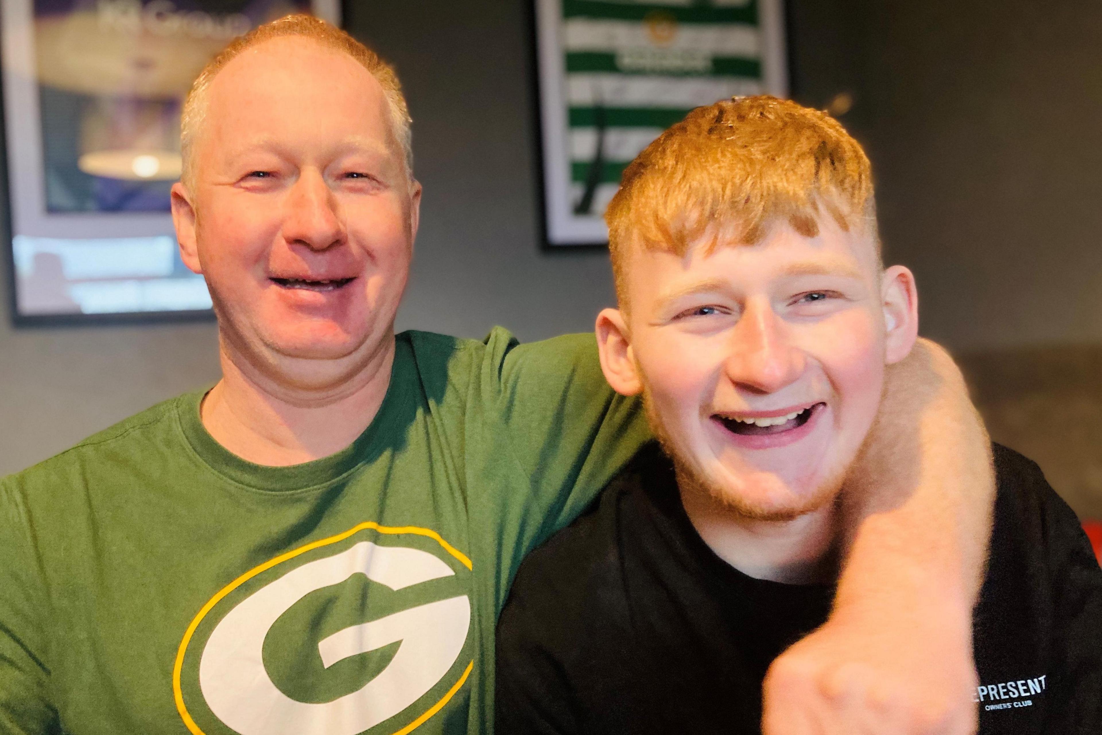 Older man smiling at camera with green T-shirt on with a large letter G on it it white with a yellow trim, with his arm round a smiling younger man - his son - smiling at camera and wearing a white T-shirt.