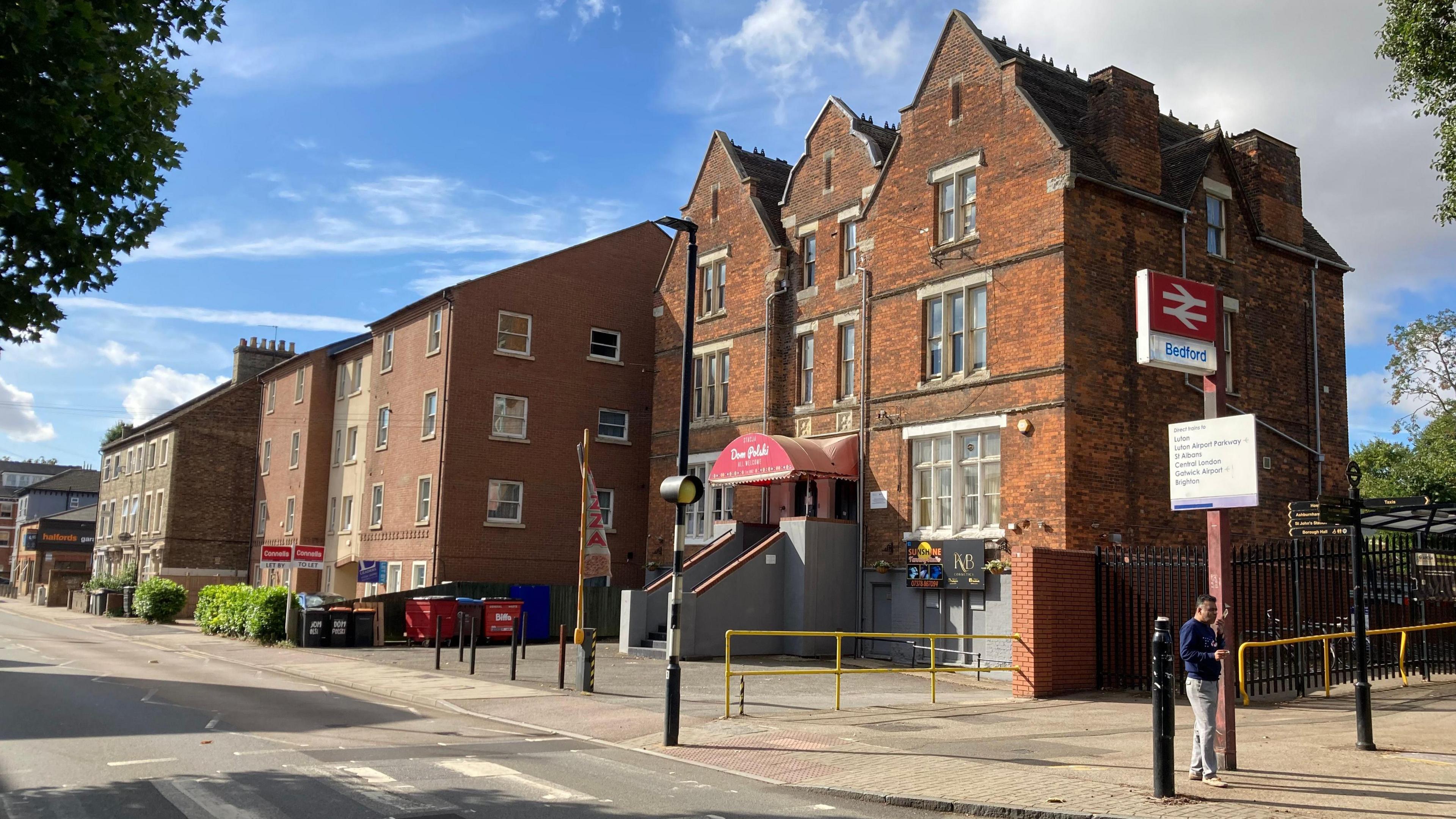 A picture of a road in Bedford, you can see the Dom Polski club which is a red brick building next to a sign for a railway station. There are also more houses down the right hand side of the road and a road crossing.