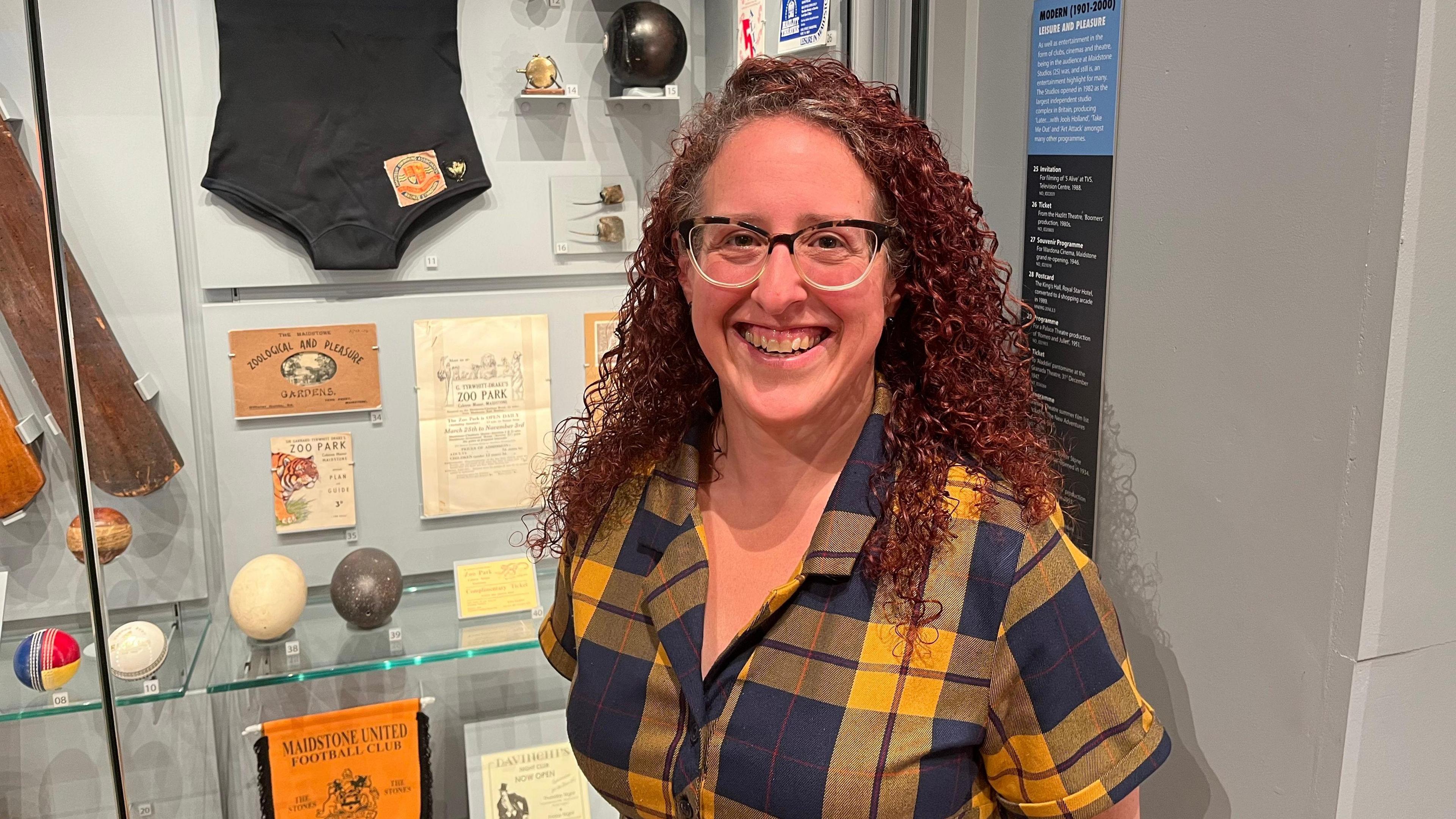 Sam Harris, Collections Manager at Maidstone Museum stands in the Oldham Gallery in front of a display case featuring sporting paraphernalia from Maidstone United and cricket teams