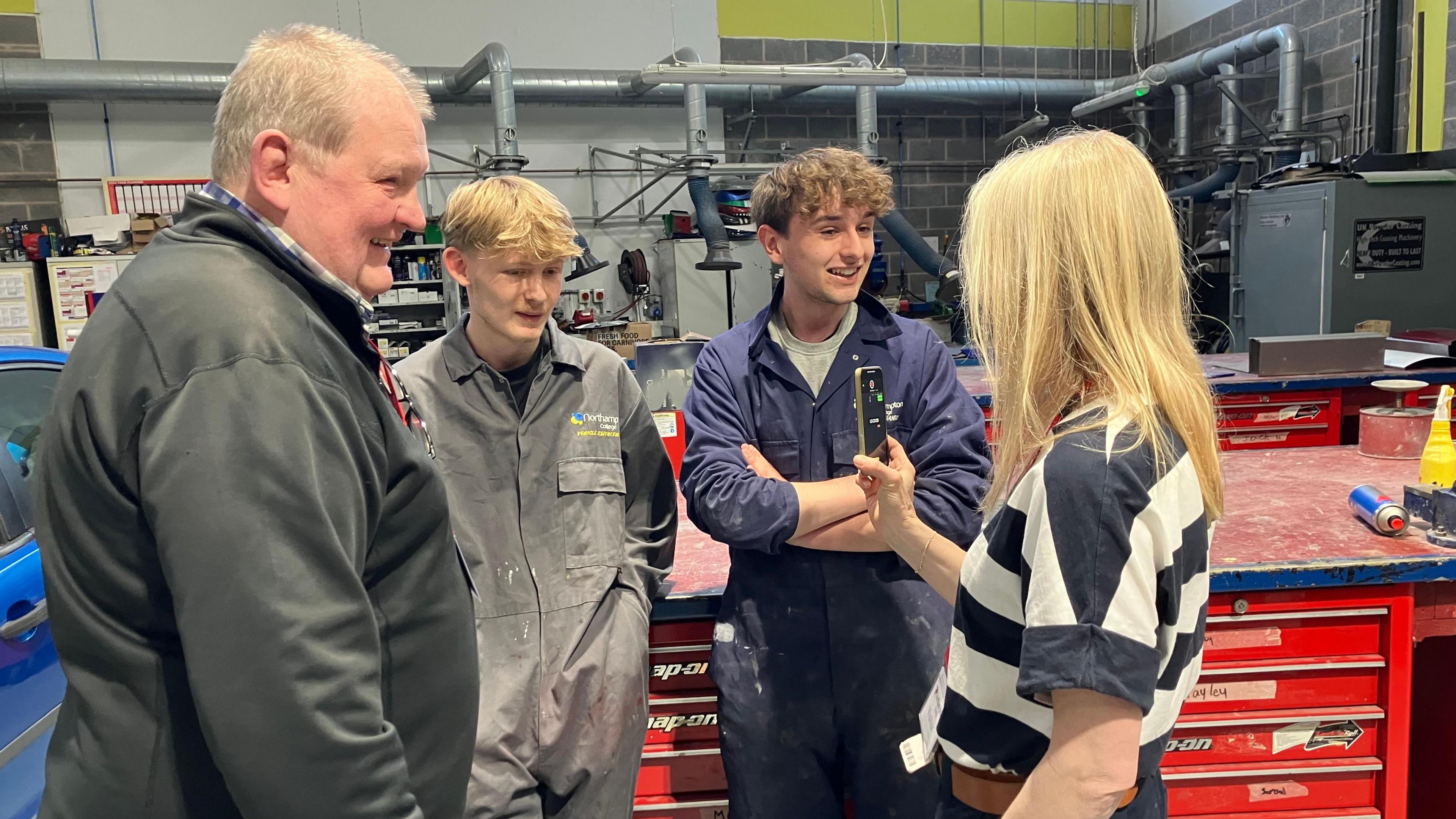 Four people in a car workshop. There are two teenage boys, one has blond hair and is wearing grey overalls, the other has brown har and is wearing blue overalls. He is talking into a microphone held by Annabel Amos with long blond hair.  :Lennie Newman with short white hair and wearing a grey top is watching.