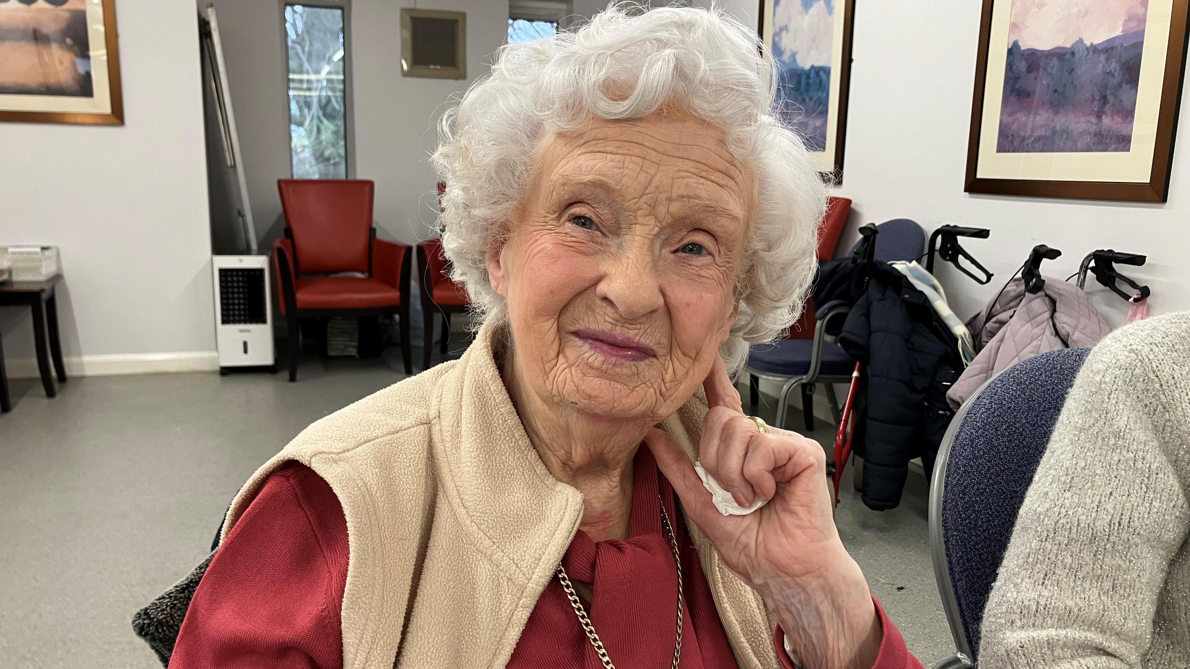 An elderly woman with white hair rests her head on her left hand while sitting down and smiling. She is wearing a crimson shirt with chain, and a cream body warmer.