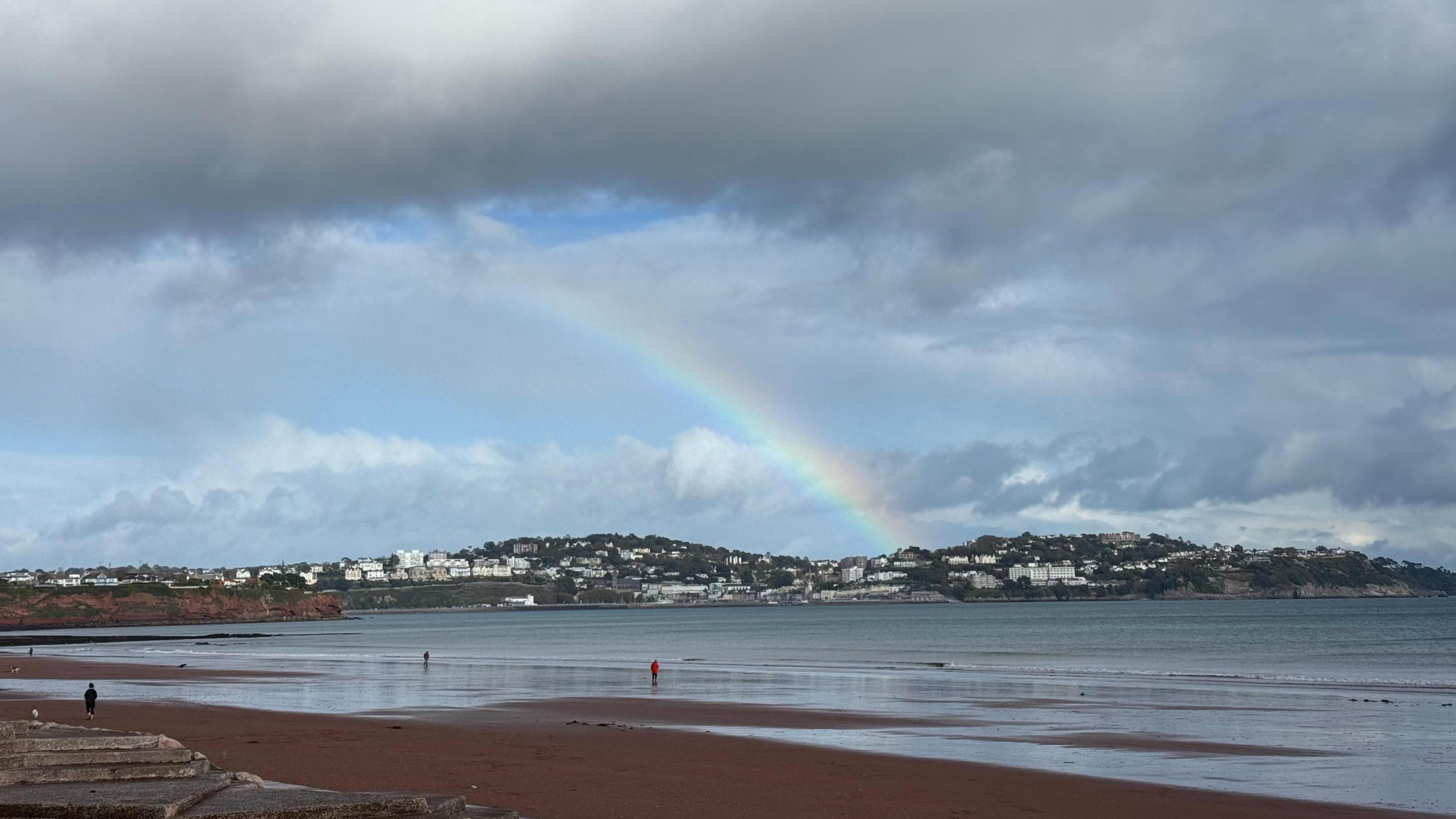 A rainbow in the background. There are grey clouds in the sky and some blue sky. In the foreground is a sandy beach. The tide is out. There are people walking along the beach. In the background there are houses on hills.