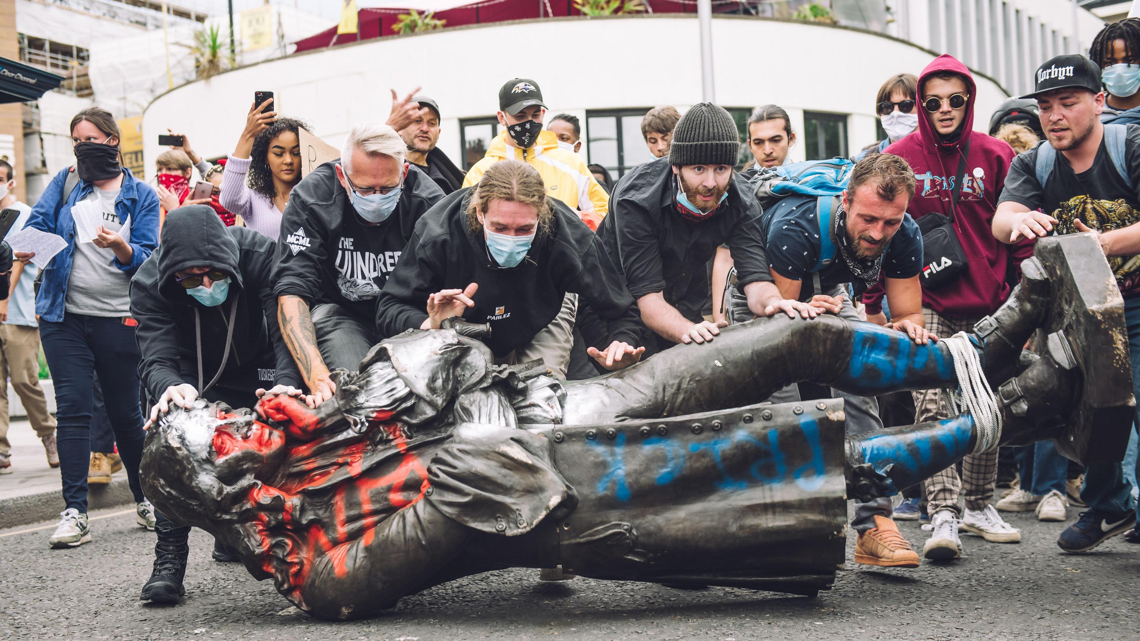 A group of people rolling the toppled Edward Colston statue through the streets of Bristol. The statue is on its side, covered in red and blue graffiti and with a rope around its feet. There is a crowd in the background following the protestors.