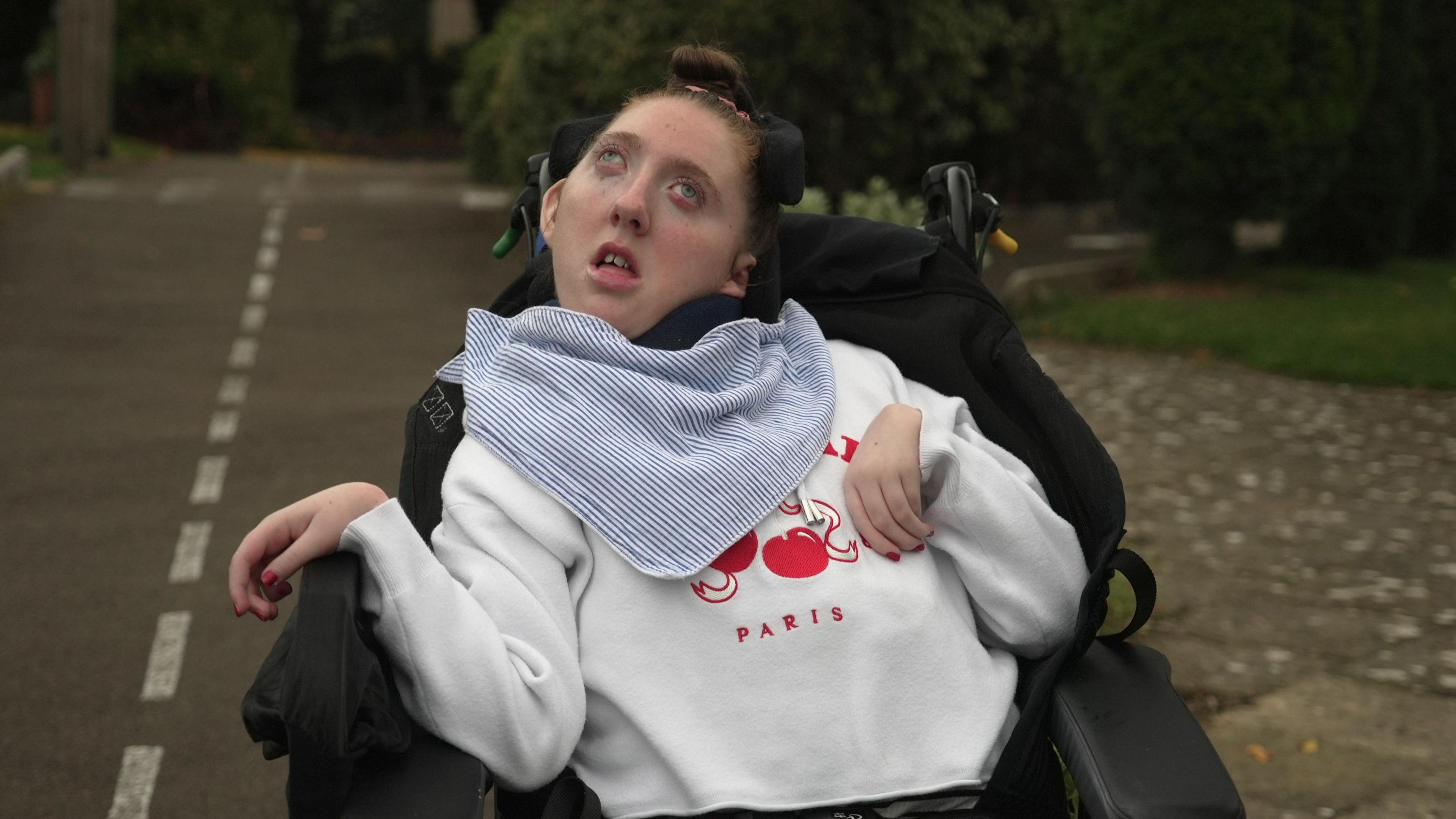 Rhi is a young woman with cerebral palsy. She is sitting in a wheelchair and has brown hair in a bun. She is wearing a white jumper.