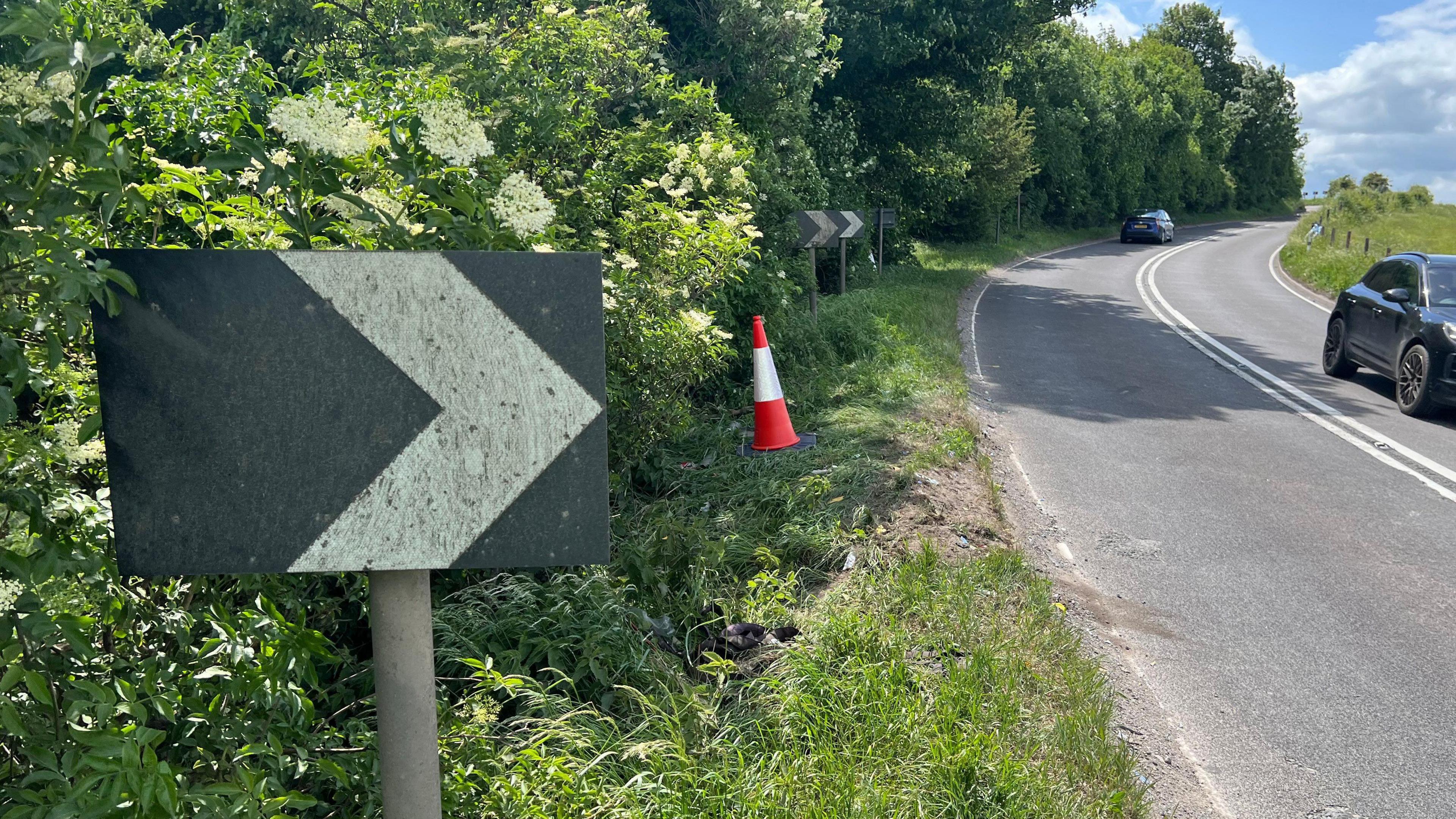 A black sign with an arrow on it on the side of the A36 in Wiltshire. The road next to the sign curves to the right. There are two cars travelling in opposite directions on the road. There is a traffic cone on the grass verge near the sign.