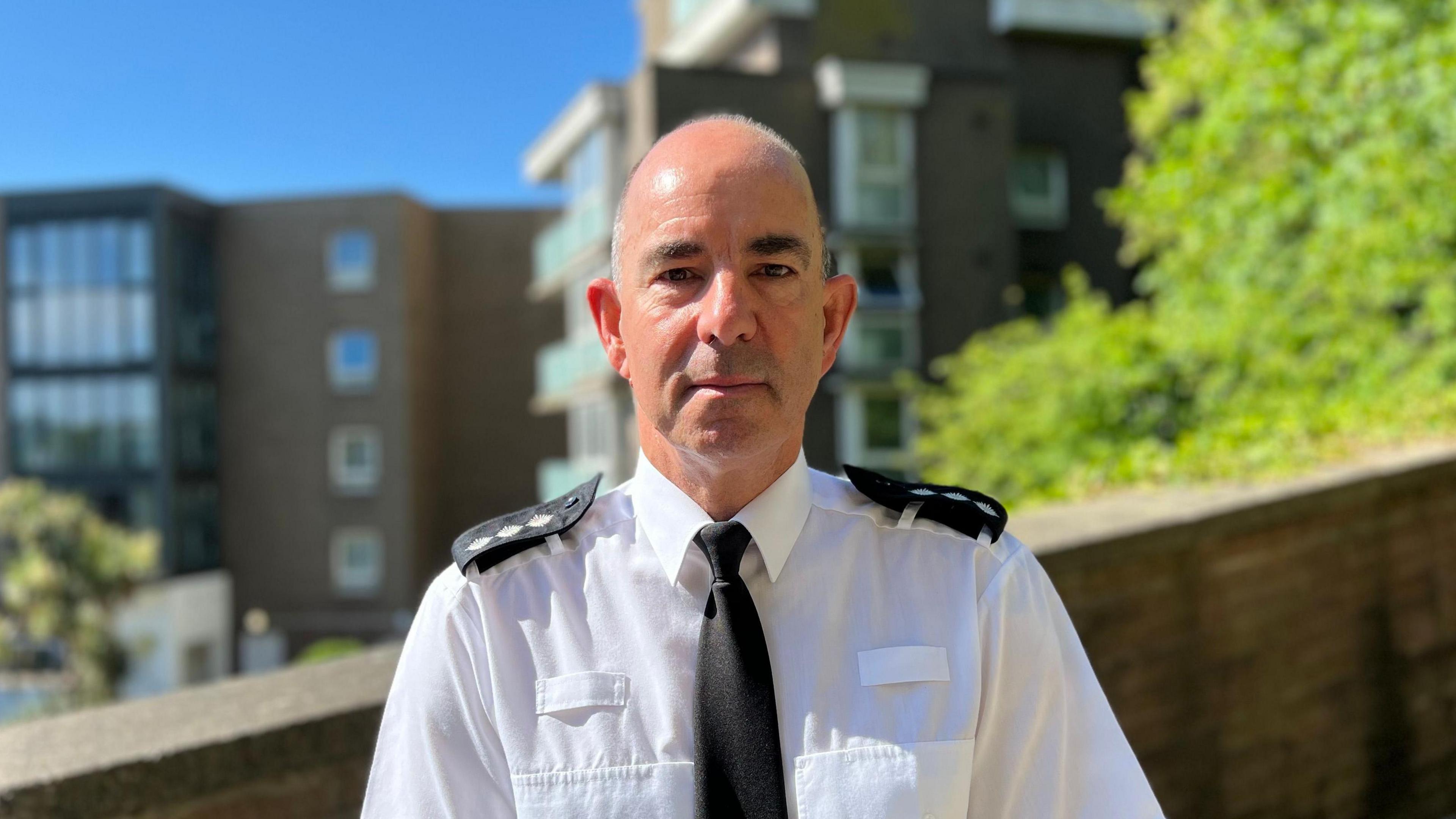 Chris Beechey is looking into the camera. He is wearing police uniform and standing in front of a brick wall. The wall is in front of a brown block of flats and some green trees.