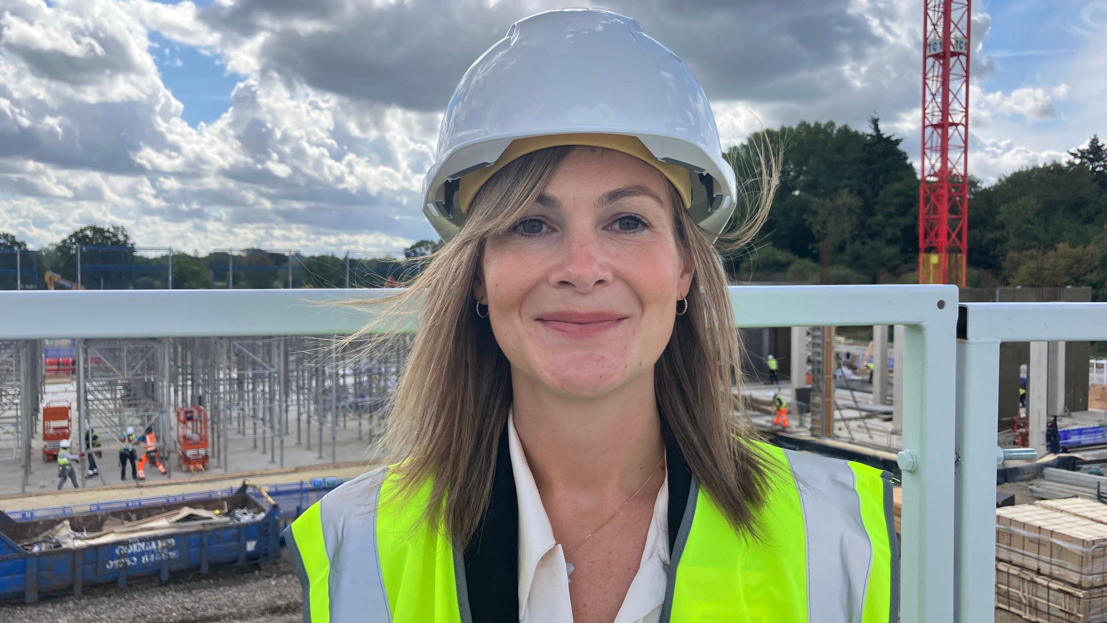 Jessica Bradford has shoulder-length blonde hair and is wearing a high visibility vest and a white hard hat. She's standing on a construction site on a sunny day and smiling at the camera.