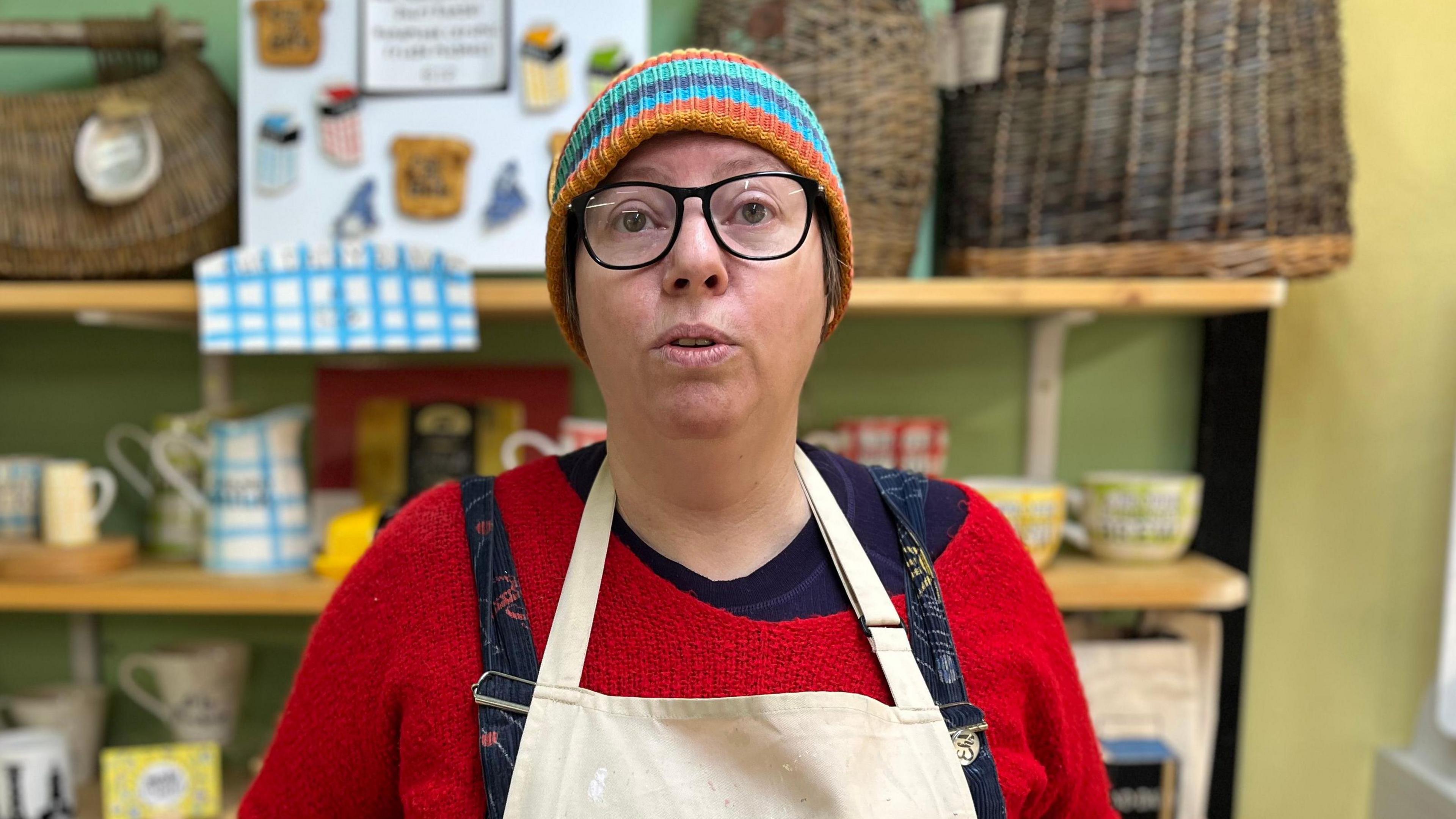 Denise McGahy - A woman wearing a multi coloured beanie hat. She's wearing an off-centre red jumper and an apron on top of dungarees. On her face are black rimmed glasses. 