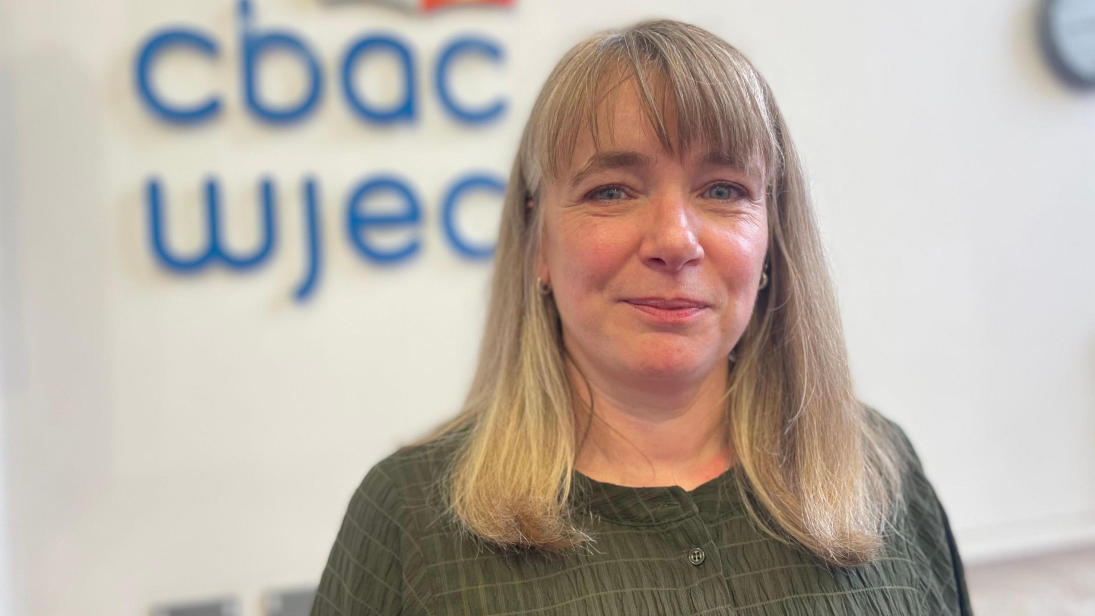 Delyth Jones o CBAC gyda gwallt golau i'w hysgwysdau yn sefyll o flaen wal gyda logo CBAC arno
