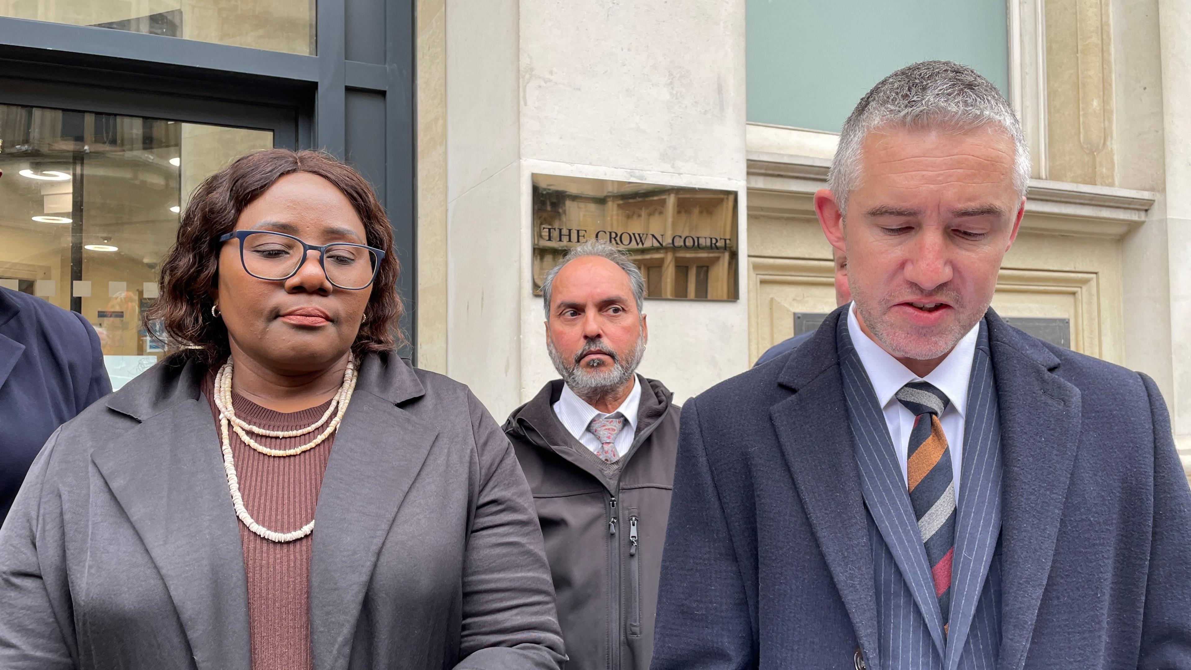 Hivaka Tjitendero standing outside Bristol Crown Court, wearing a brown shirt and black blazer, with a layered necklace and black framed glasses. She looks solemn and is looking towards the ground. She is joined by Det Supt Mike Buck, who is wearing a blue suit and striped tie. He is looking down as he reads a statement on behalf of the family.