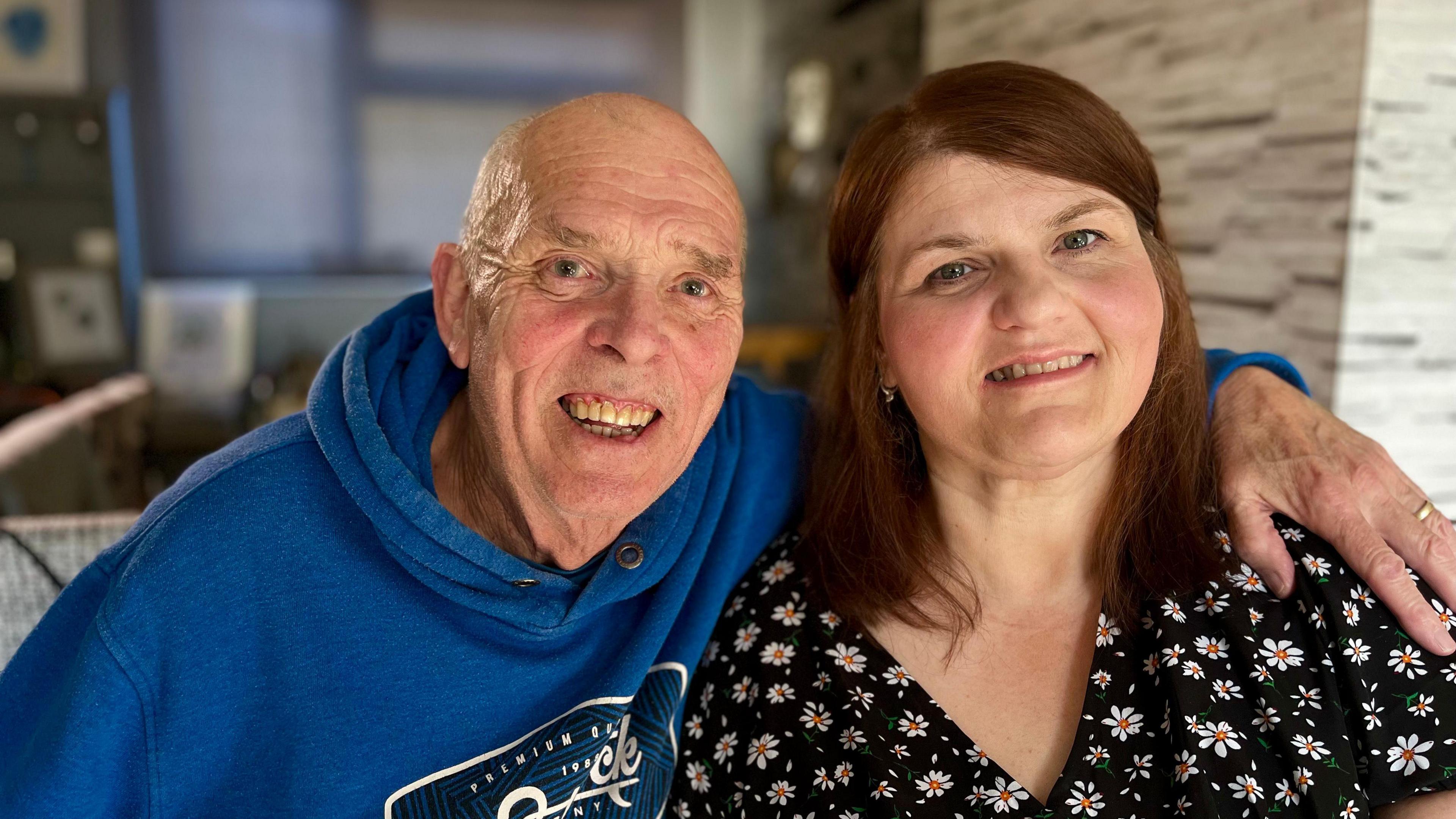 Dave Holmes and Kelly Holmes sitting side by side in their home and smiling at the camera. Dave is sat on the left and has his arm over Kelly's shoulders in what looks to be a comforting gesture. He wears a blue hoodie and appears to have no hair. Kelly is wearing a black blouse with white flowers on which have a dot of yellow - resembling the pollen - in the centre. She has brown-red - almost chestnut colour - shoulder length hair which has been tucked behind her ears.