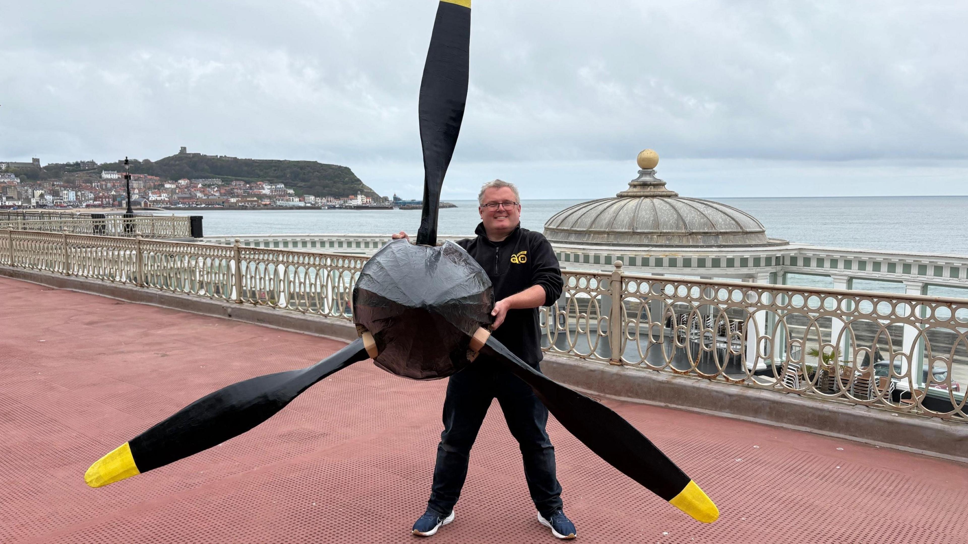A man dressed in black on the seafront with the propellor from a replica Mk1 Spitfire in his hands