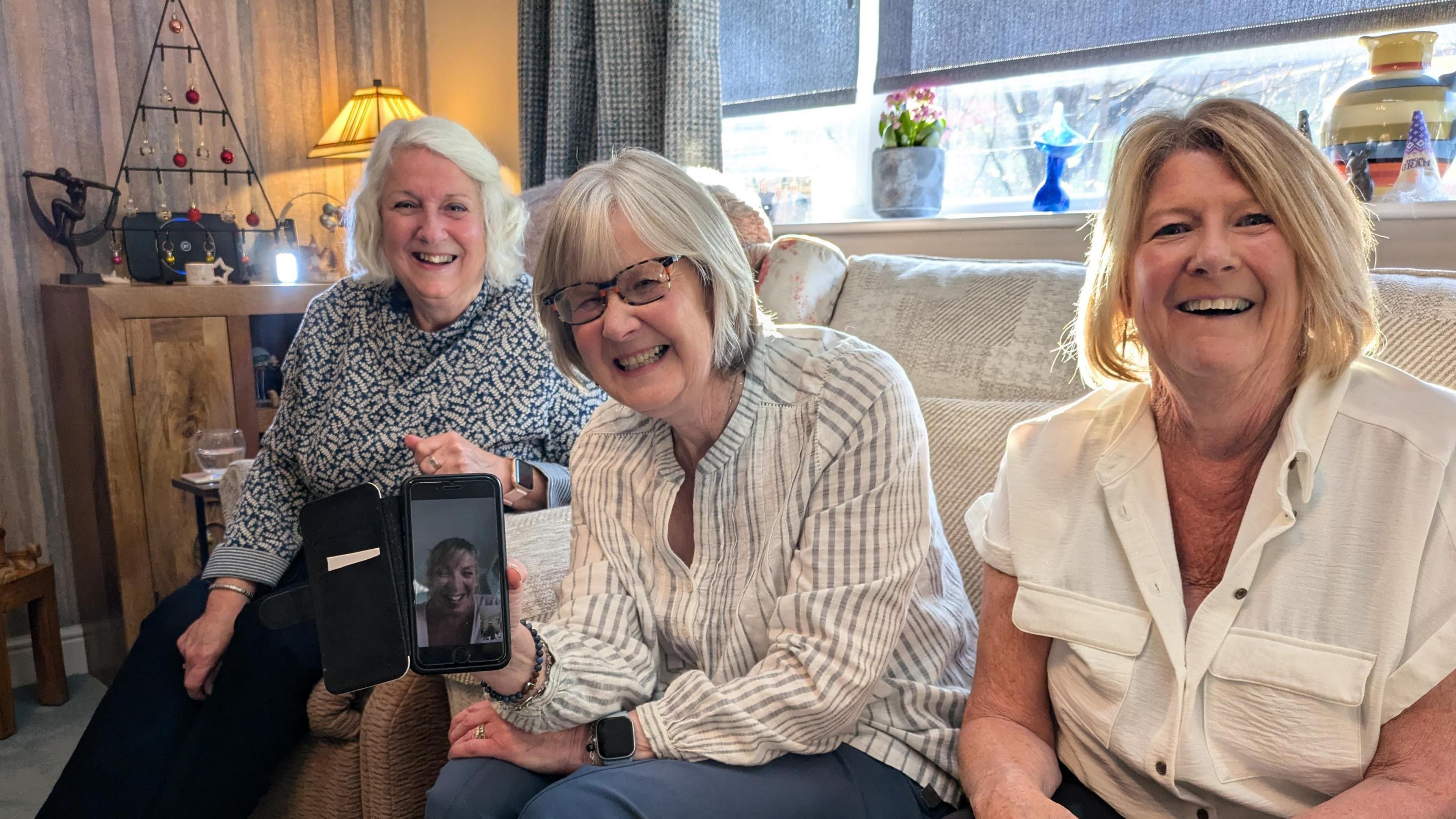 Three women sat on a sofa in a living room. All are smiling at the camera. The middle one of the three holds aloft her phone, on which their friend is on camera via Zoom.