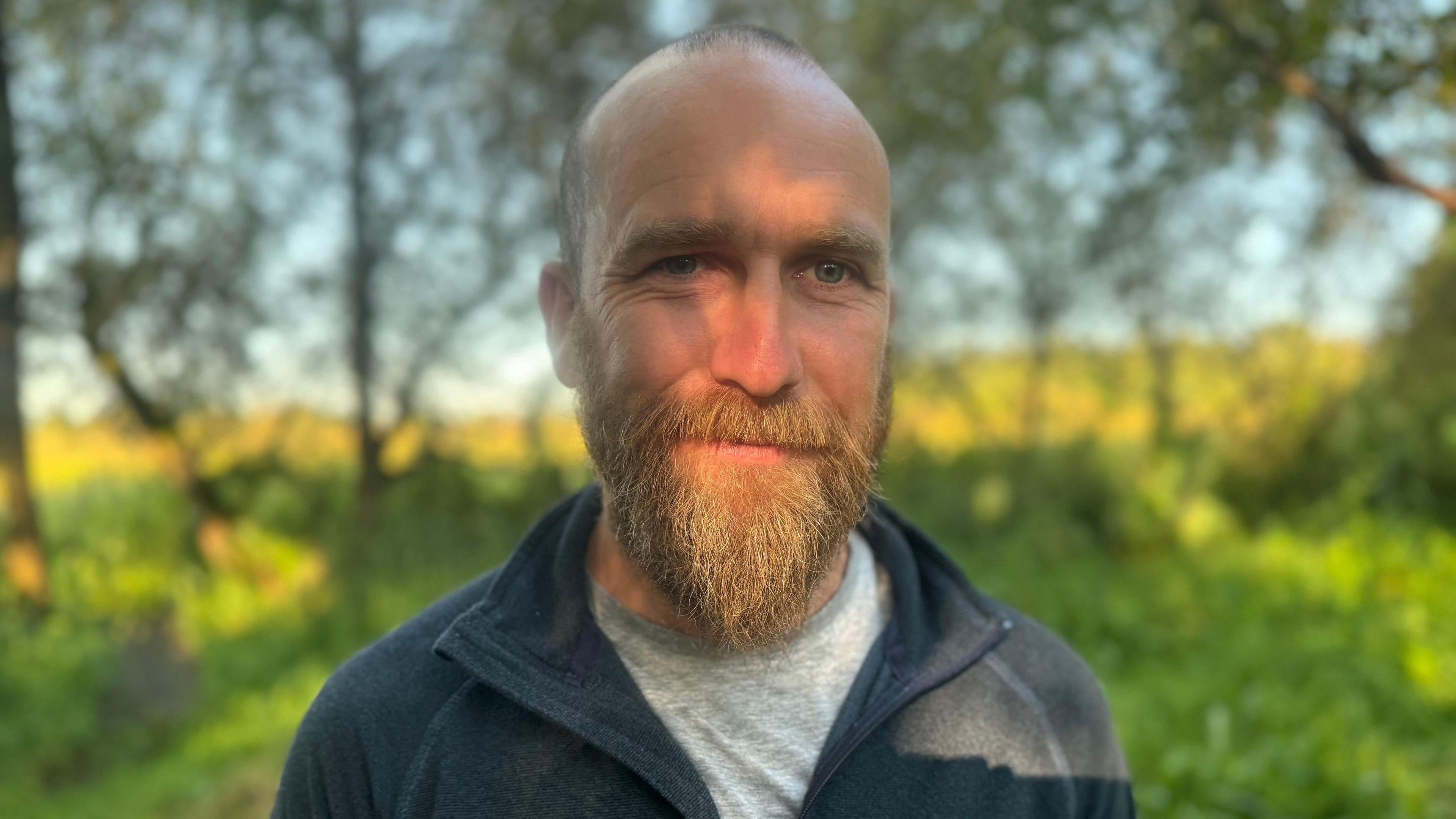 A man with a slight smile looking directly into the camera. He is almost bald with a short but thick light-coloured beard. He is wearing a dark great fleece and grey t-shirt. He is standing in front of a row of trees in the countryside during the daytime.