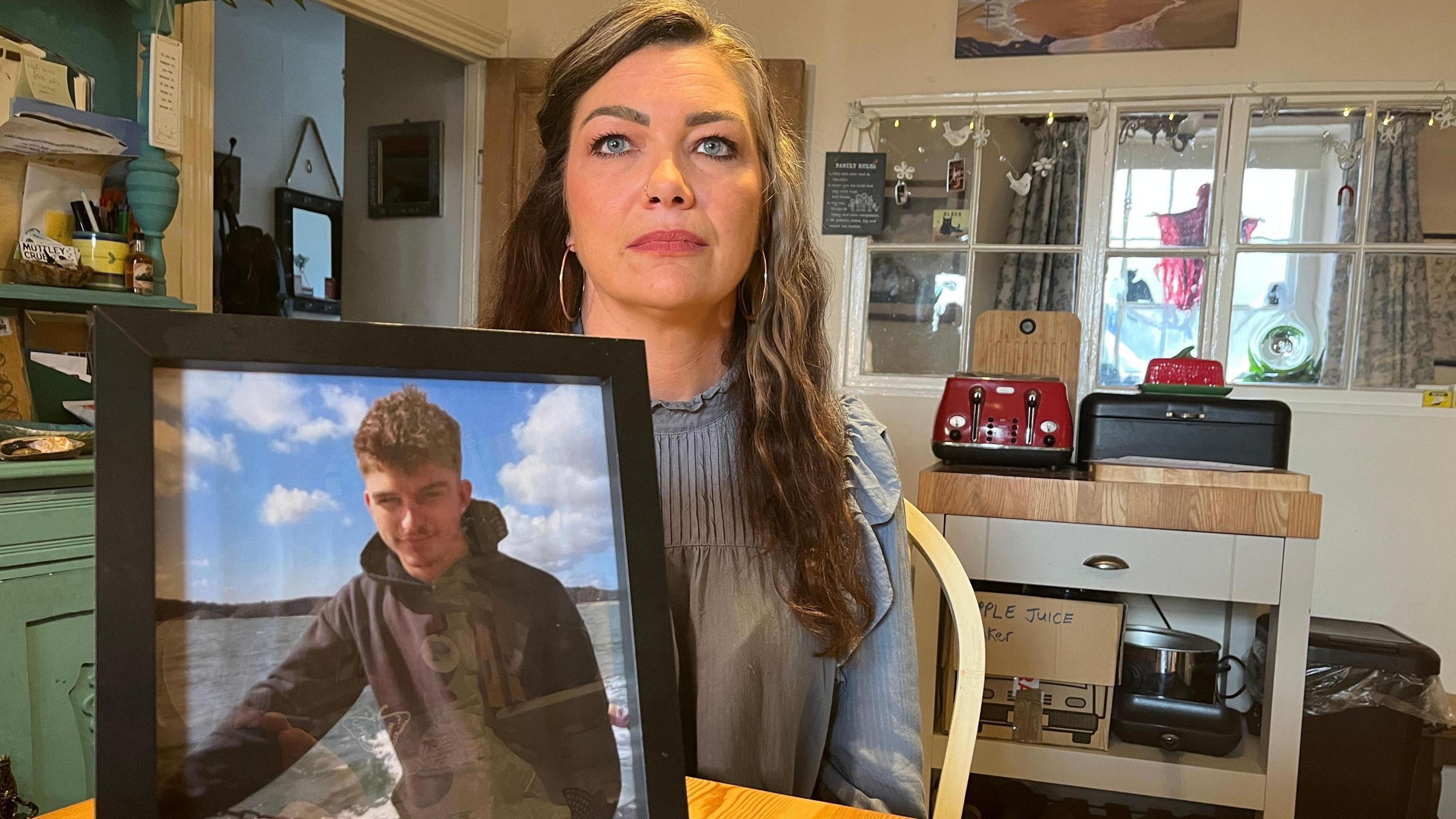 A woman with long dark hair, wearing a blue top and sat in her home, is holding up a picture of a young man with dark hair and a black hoody
