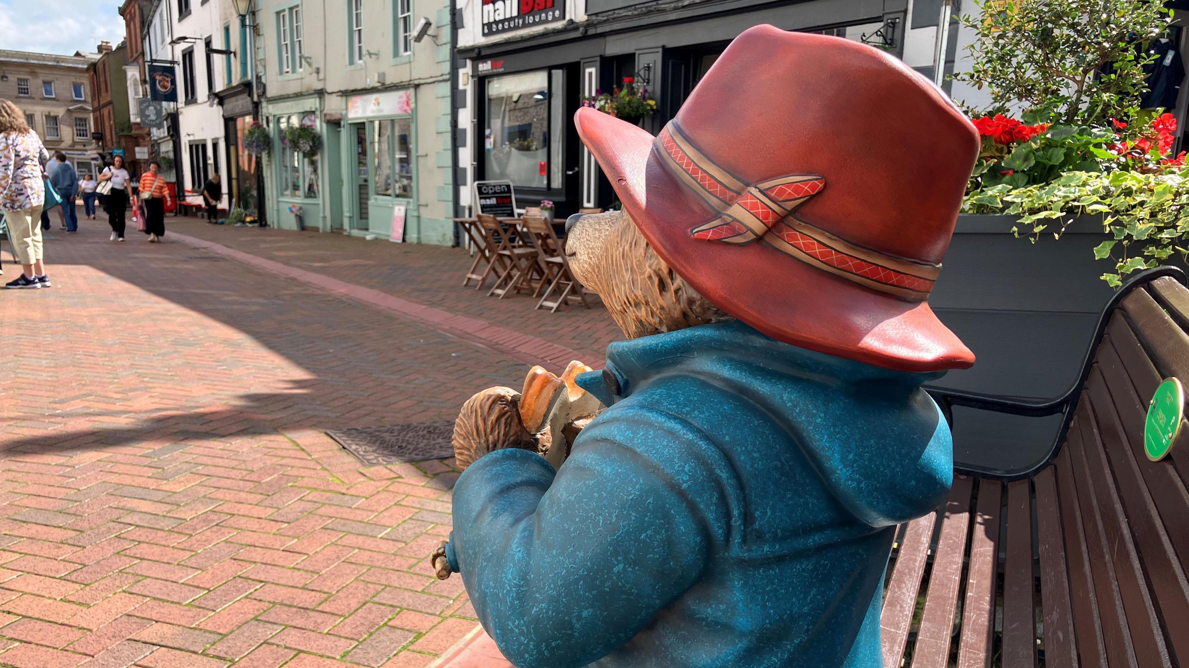 The Paddington Bear statue in Penrith pictured from behind its head, looking out to the town. The bear is sitting on a wooden bench holding a marmalade sandwich. He is wearing a red had and blue raincoat. The bench is next to a flower planter and looks out to the shops.
