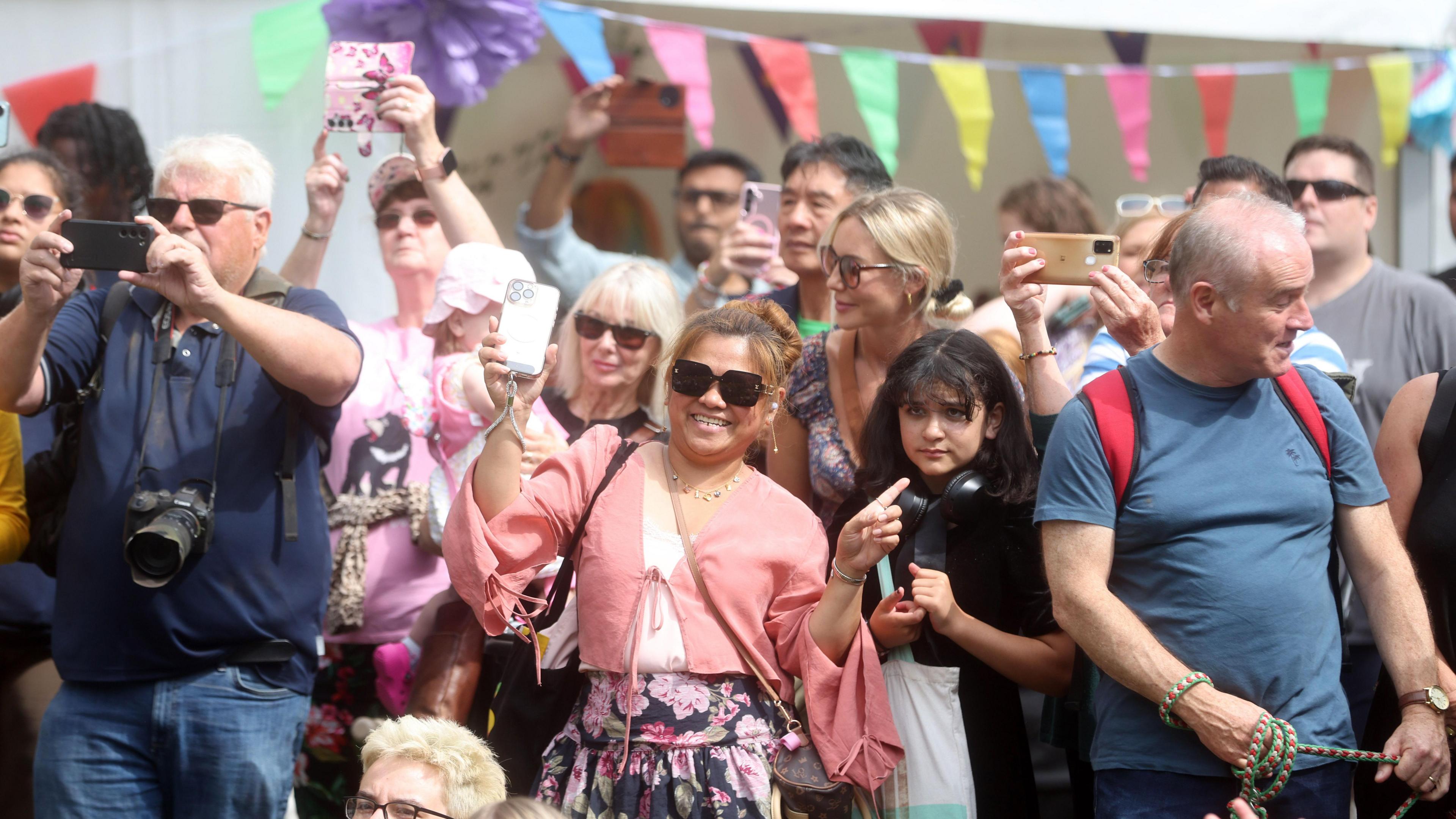 A crowd are standing together. Some people have phones in the air taking pictures. They are smiling and wearing sunglasses. 