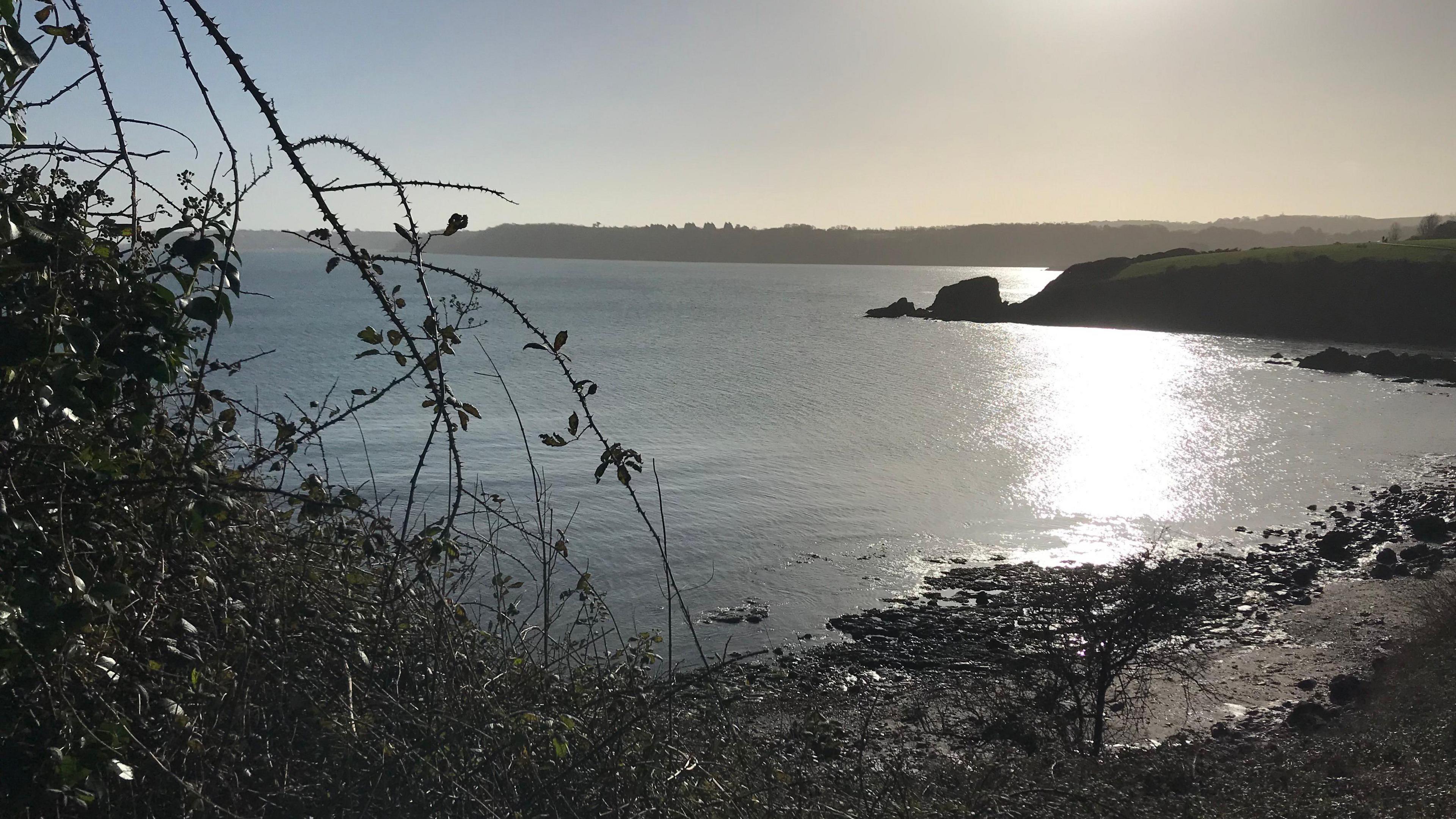 A cove is seen with the sun shimmering on the sea and a rock formation in the background