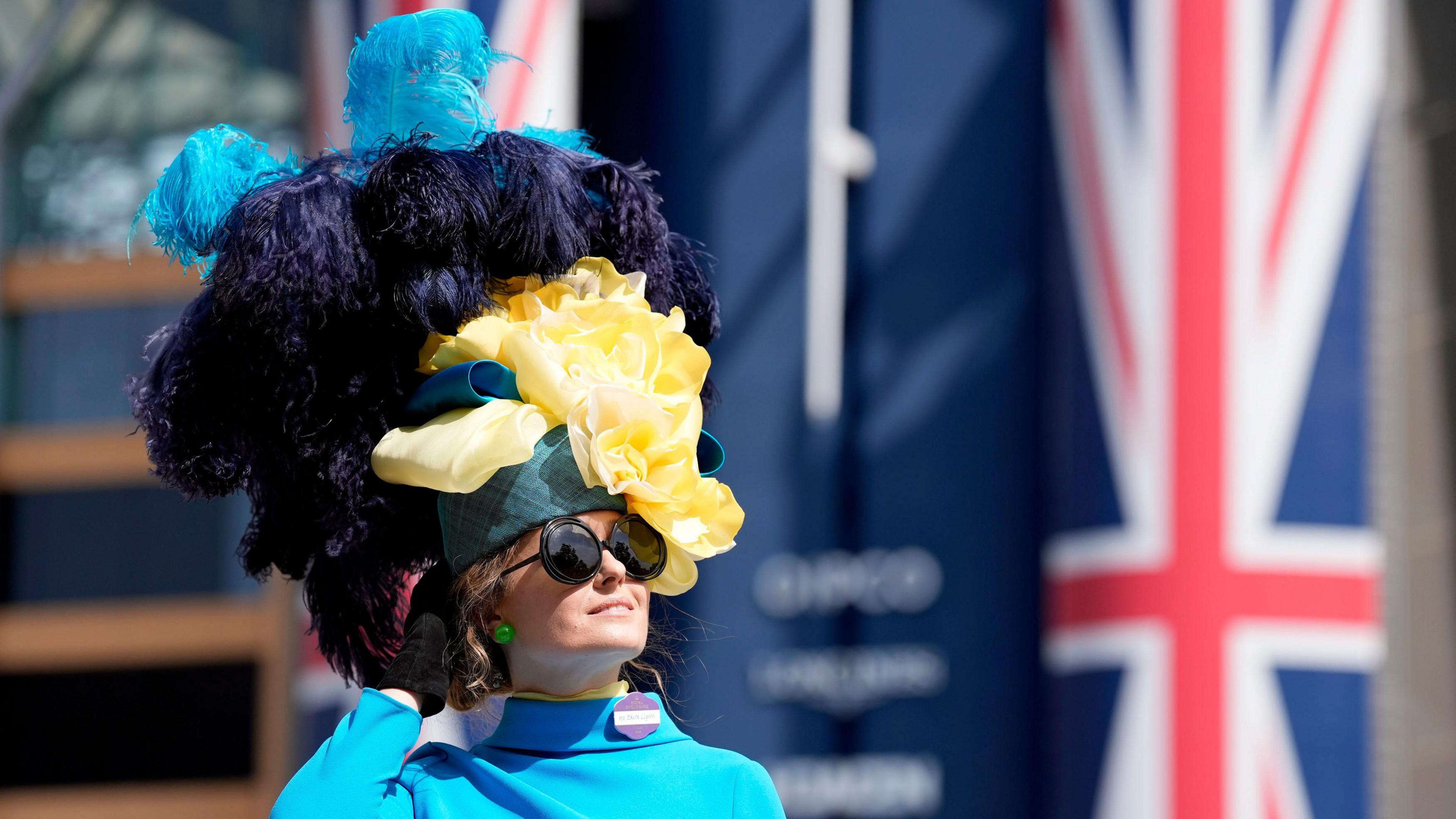 A woman wearing an elaborate fascinator, which is very tall and contains large feathers, which are light blue and dark blue. 