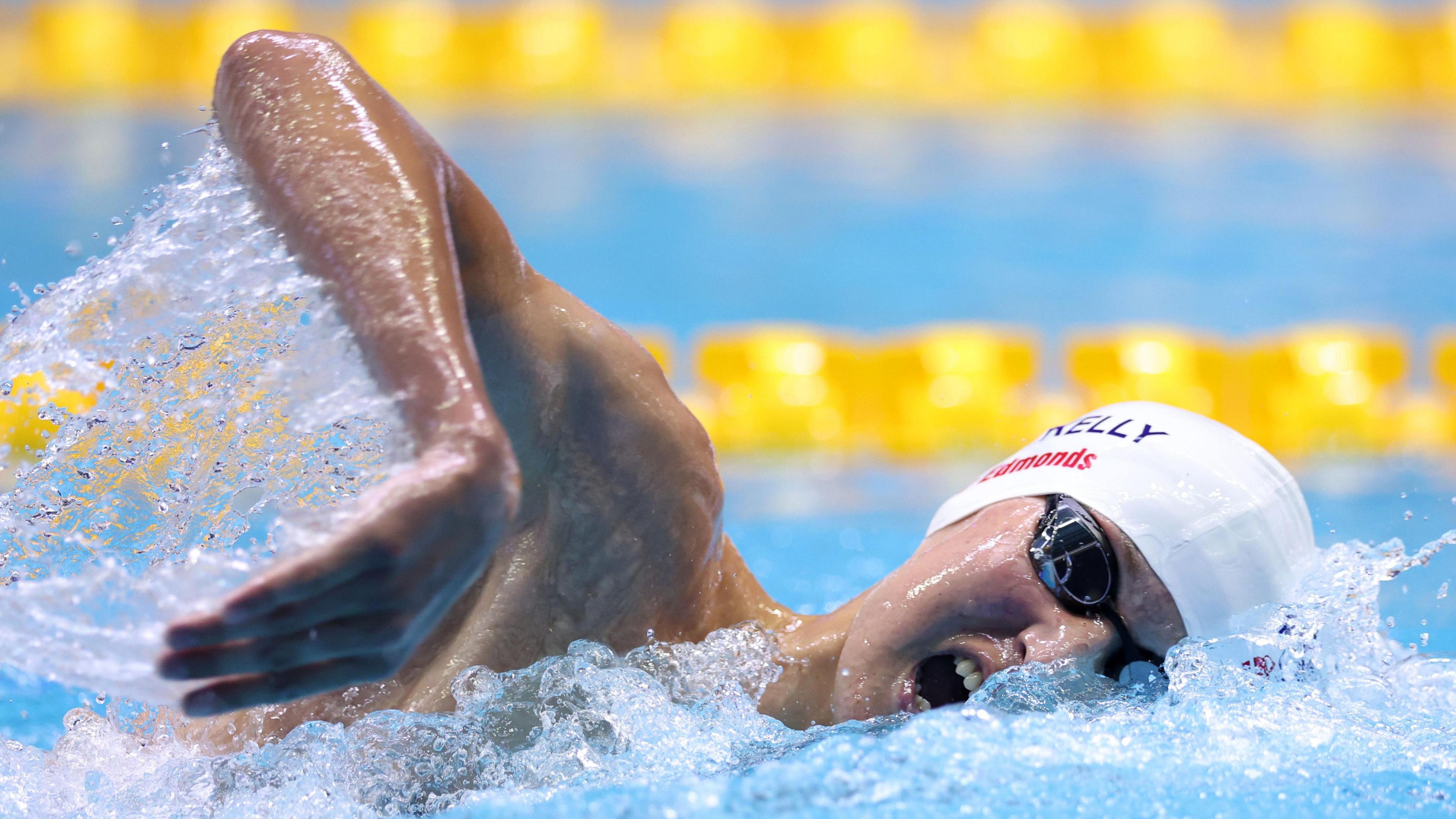 A boy swimming in a pool. He is captured moving with one arm coming up out of the water in a stroke. Half his face is underwater, but his mouth is open. He is wearing a white swimming cap and black goggles.
