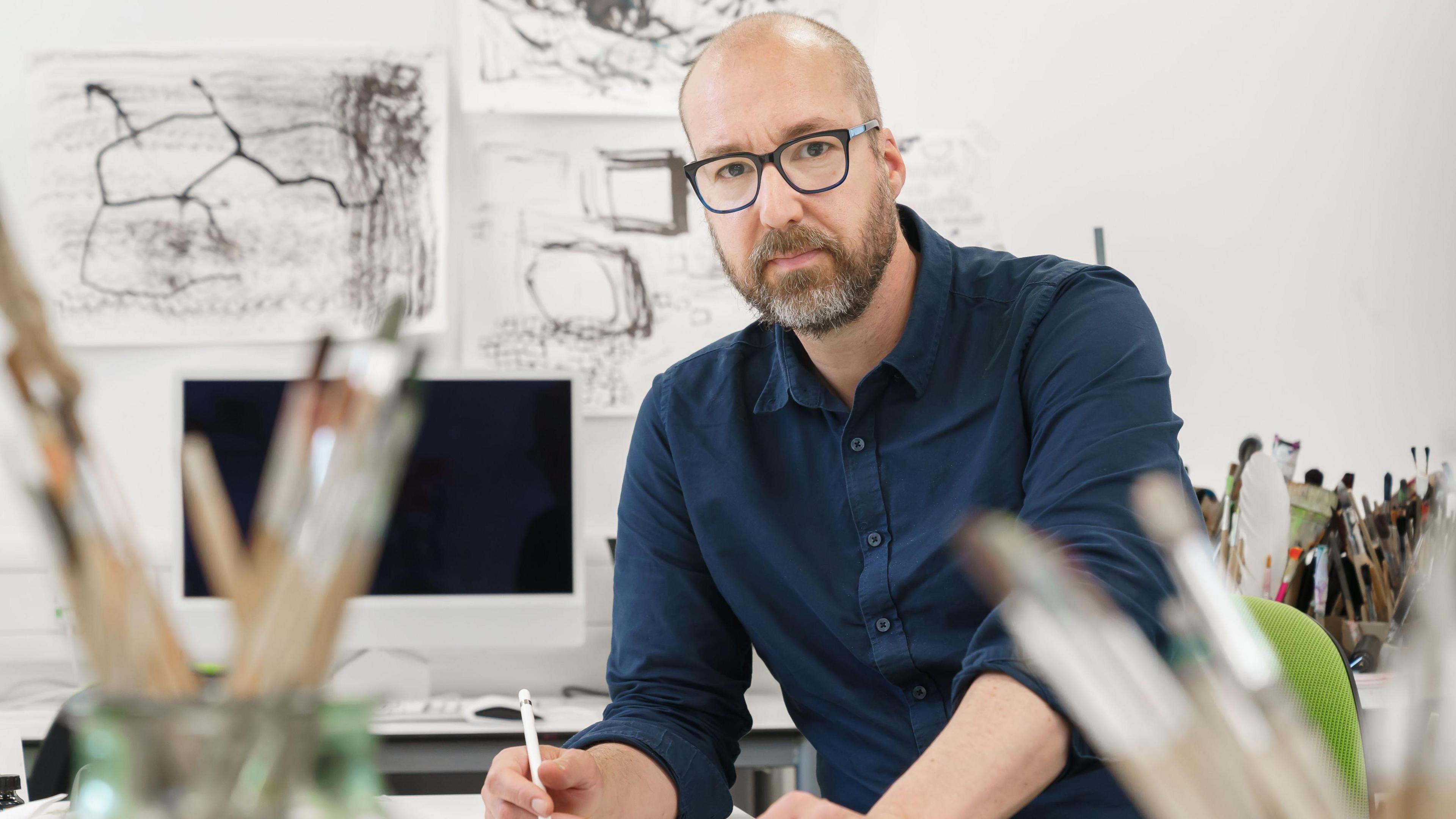 Nick Lewis looks at the camera while posing with a pencil in his hand. He is wearing a navy shirt and glasses, is bald with a dark and grey beard. Behind him is a computer and several charcoal sketches of shapes on the wall.