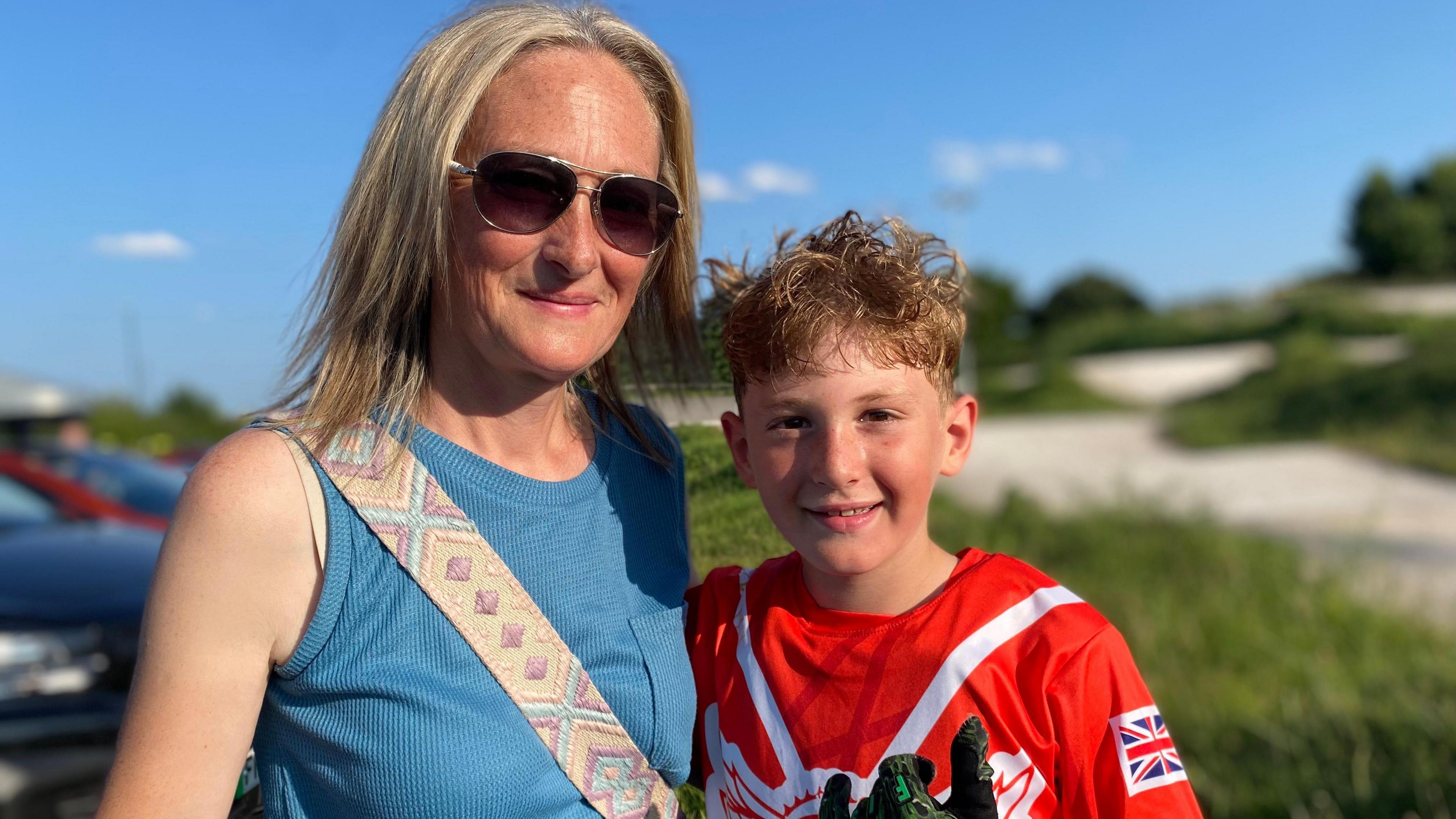 Woman wearing sunglasses and a blue top alongside a boy wearing a red top and biker gloves.