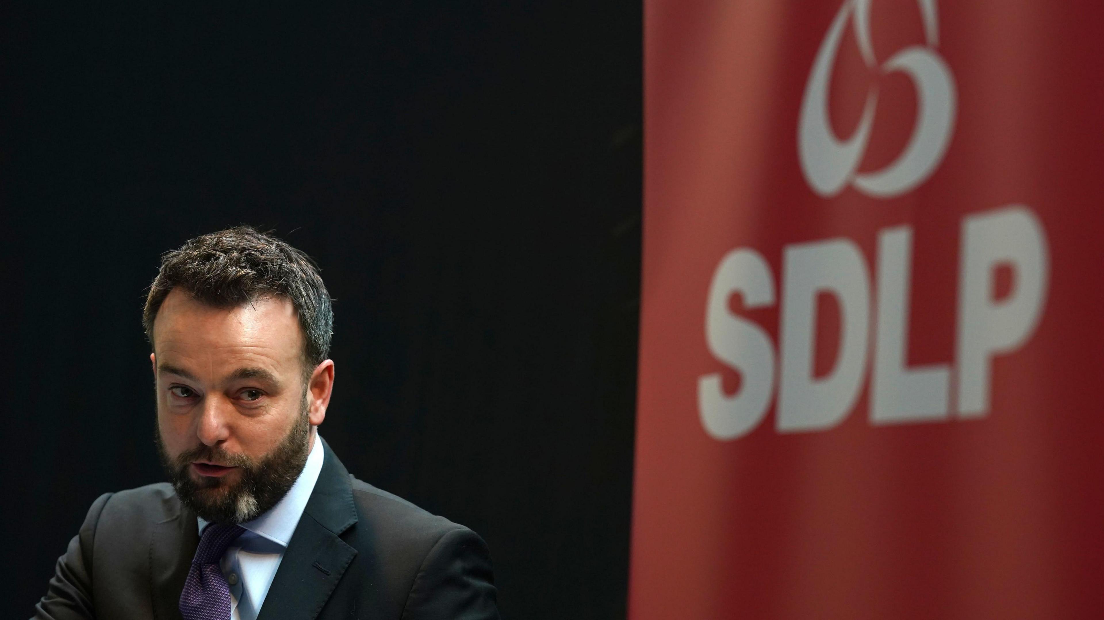 SDLP leader Colum Eastwood speaking at an event. He is wearing a dark suit and tie. Behind him is a red SDLP banner. He has dark short hair and a dark beard. 