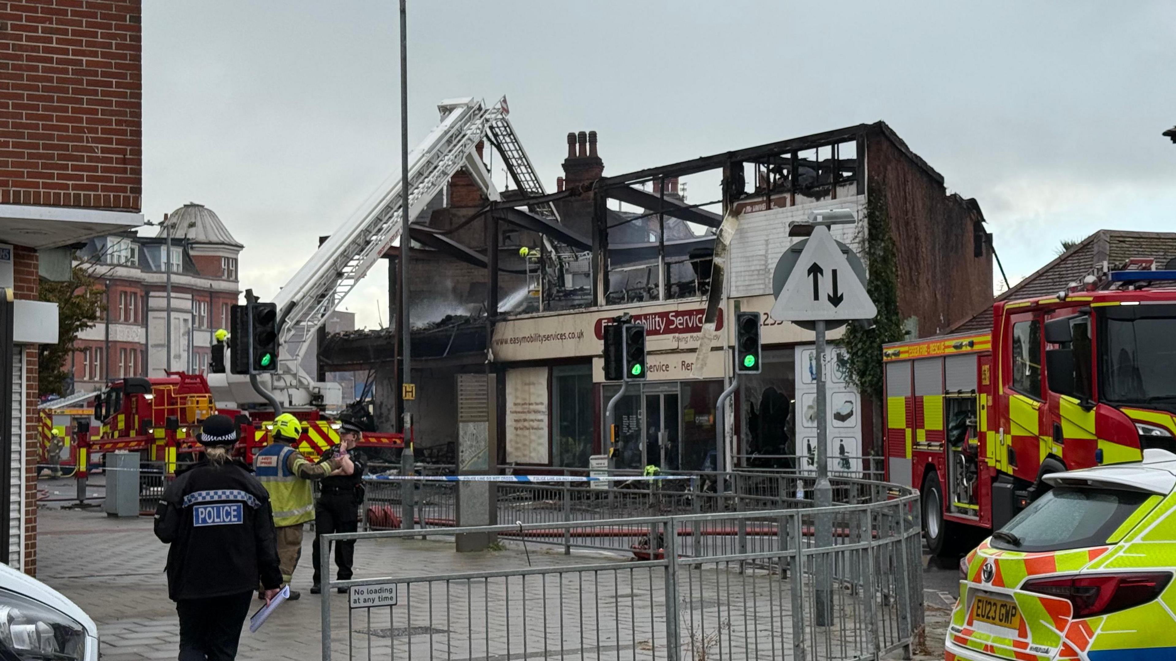 Fire crews and police on the scene this morning. Hoses are being used to spray down a brick building with missing windows, a missing roof and charred walls. The sign on the front of the building is pale yellow and reads 'easy mobility services'.